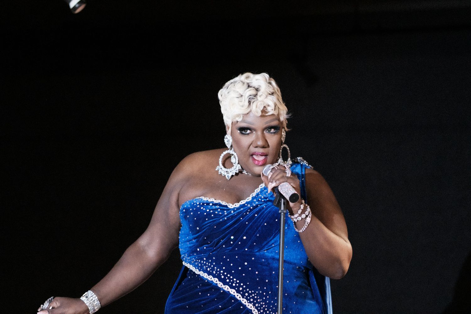 Performer in sparkling blue dress singing passionately on stage.