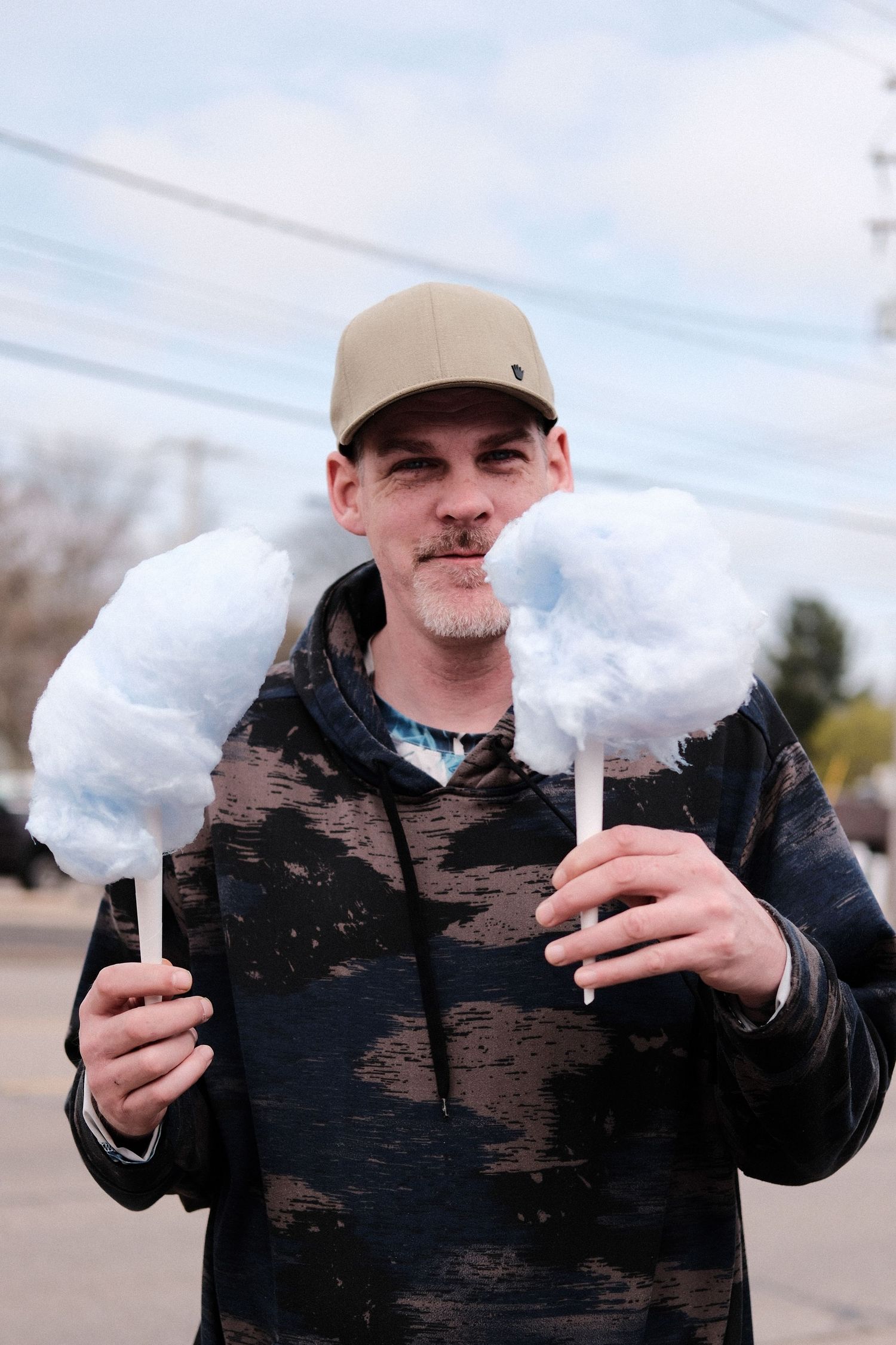 Person holding two large fluffy cotton candy sticks while wearing a baseball cap and dark jacket.