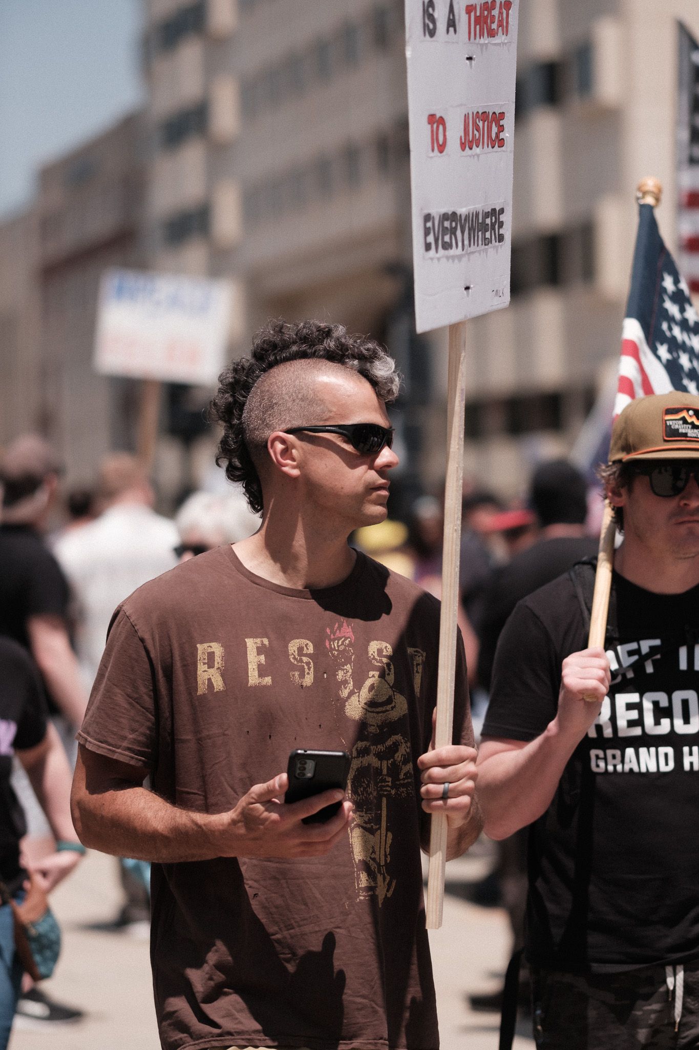 Protestor in brown t-shirt holding sign while checking phone during demonstration.