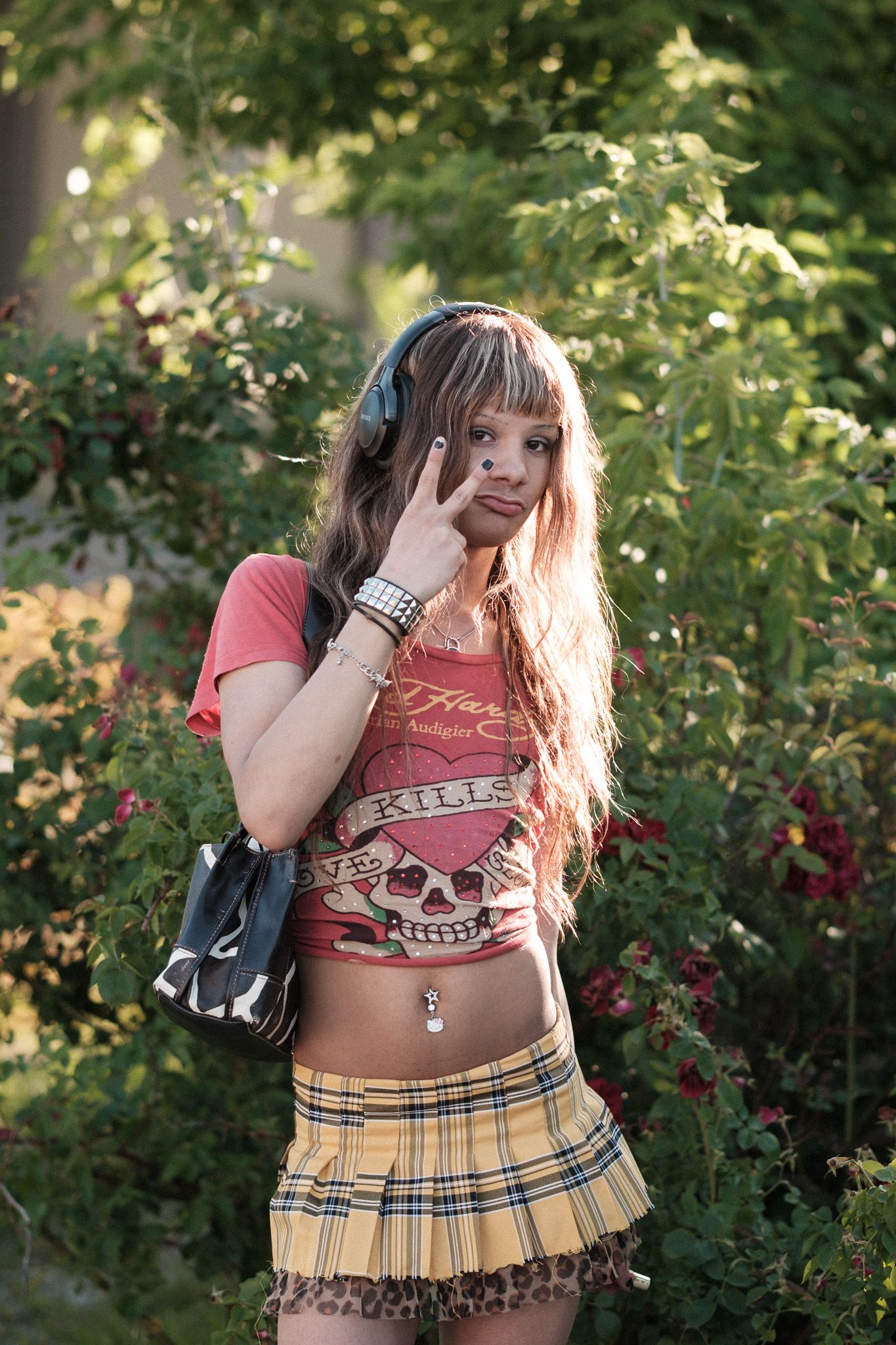 Young woman in plaid skirt and graphic tee poses in front of leafy green foliage.