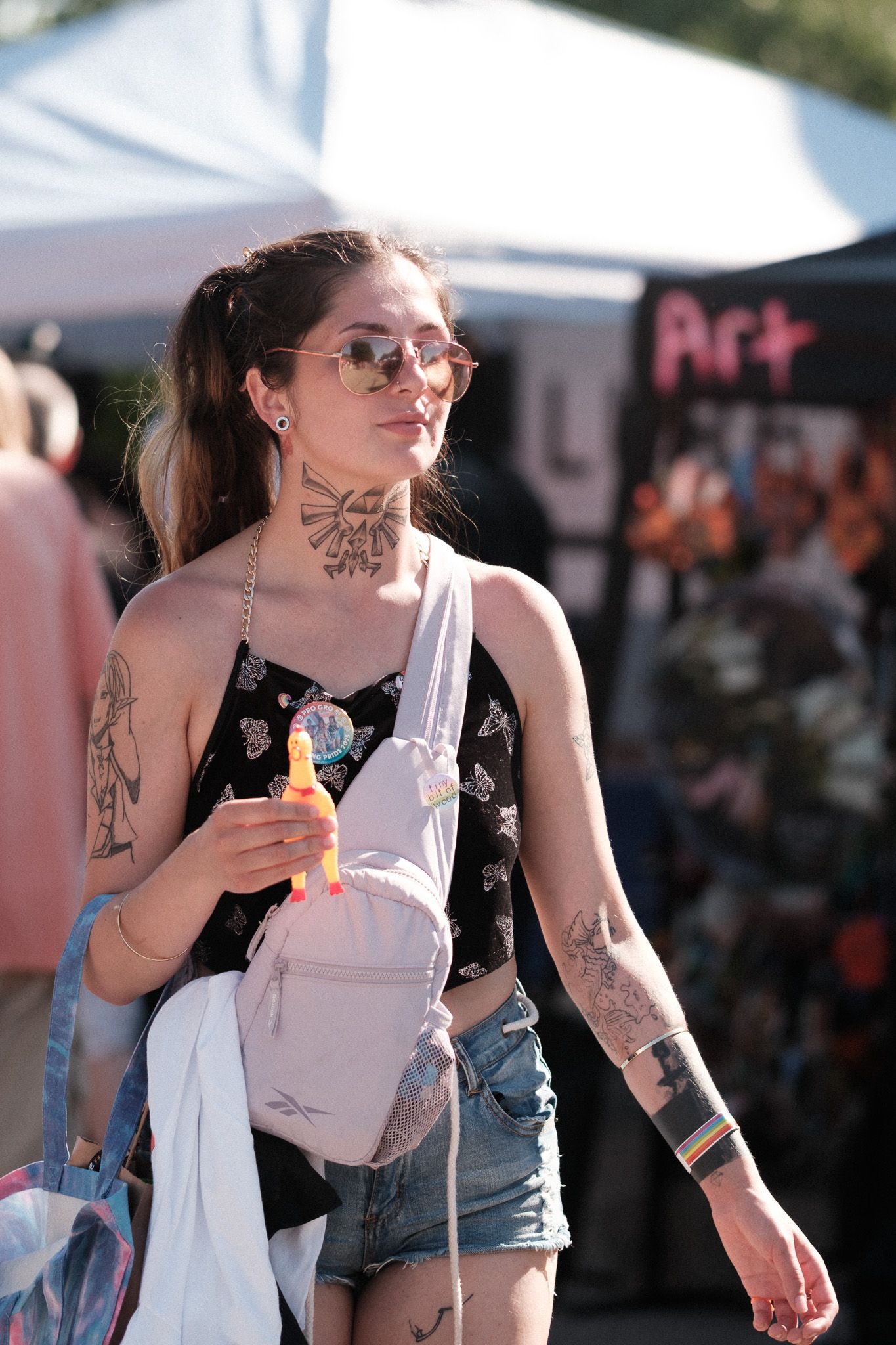 Tattooed festival goer in sunglasses and denim shorts carries a white backpack at an outdoor event.