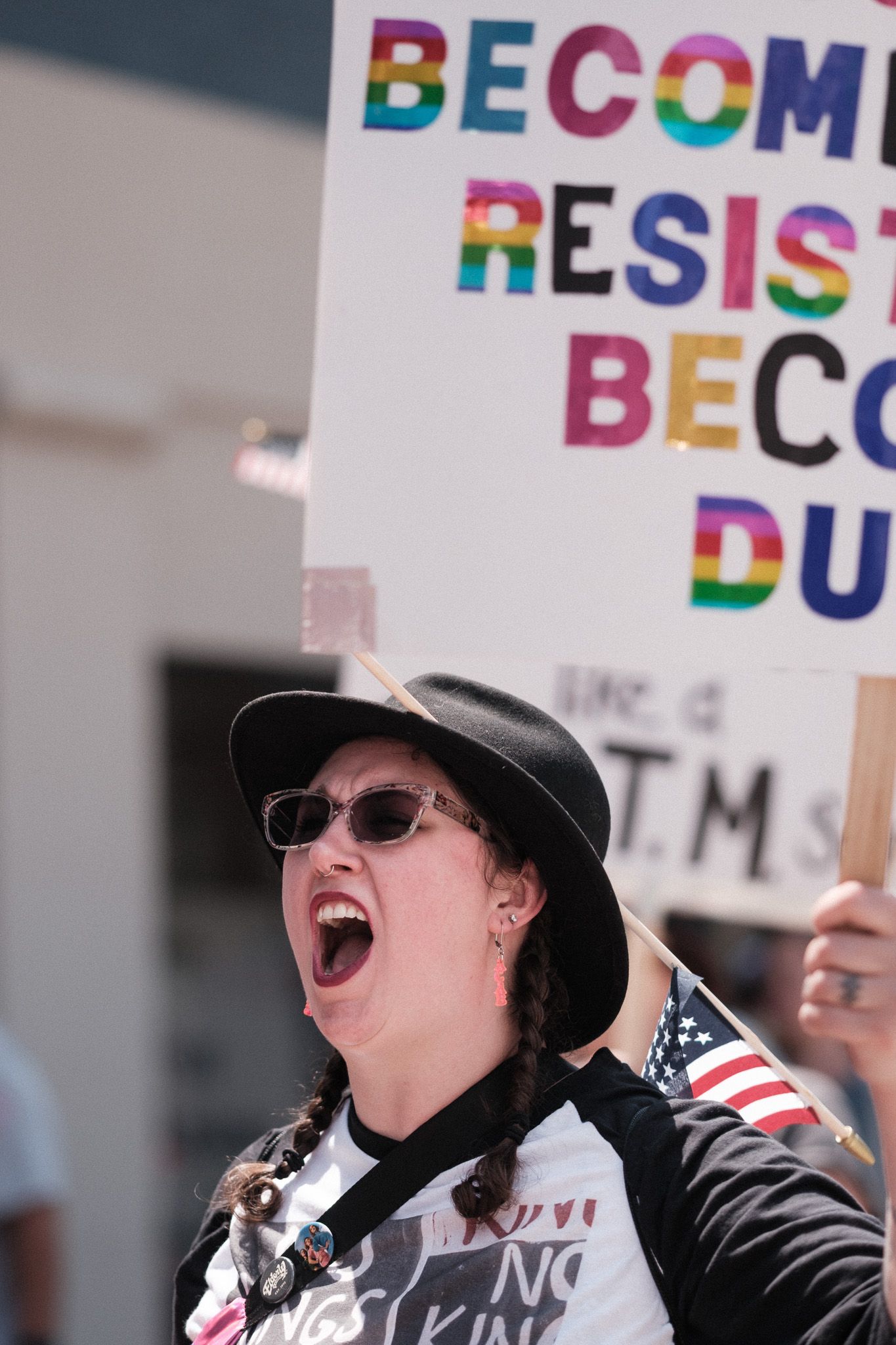 A demonstrator in a hat and sunglasses shouts while holding a colorful protest sign.