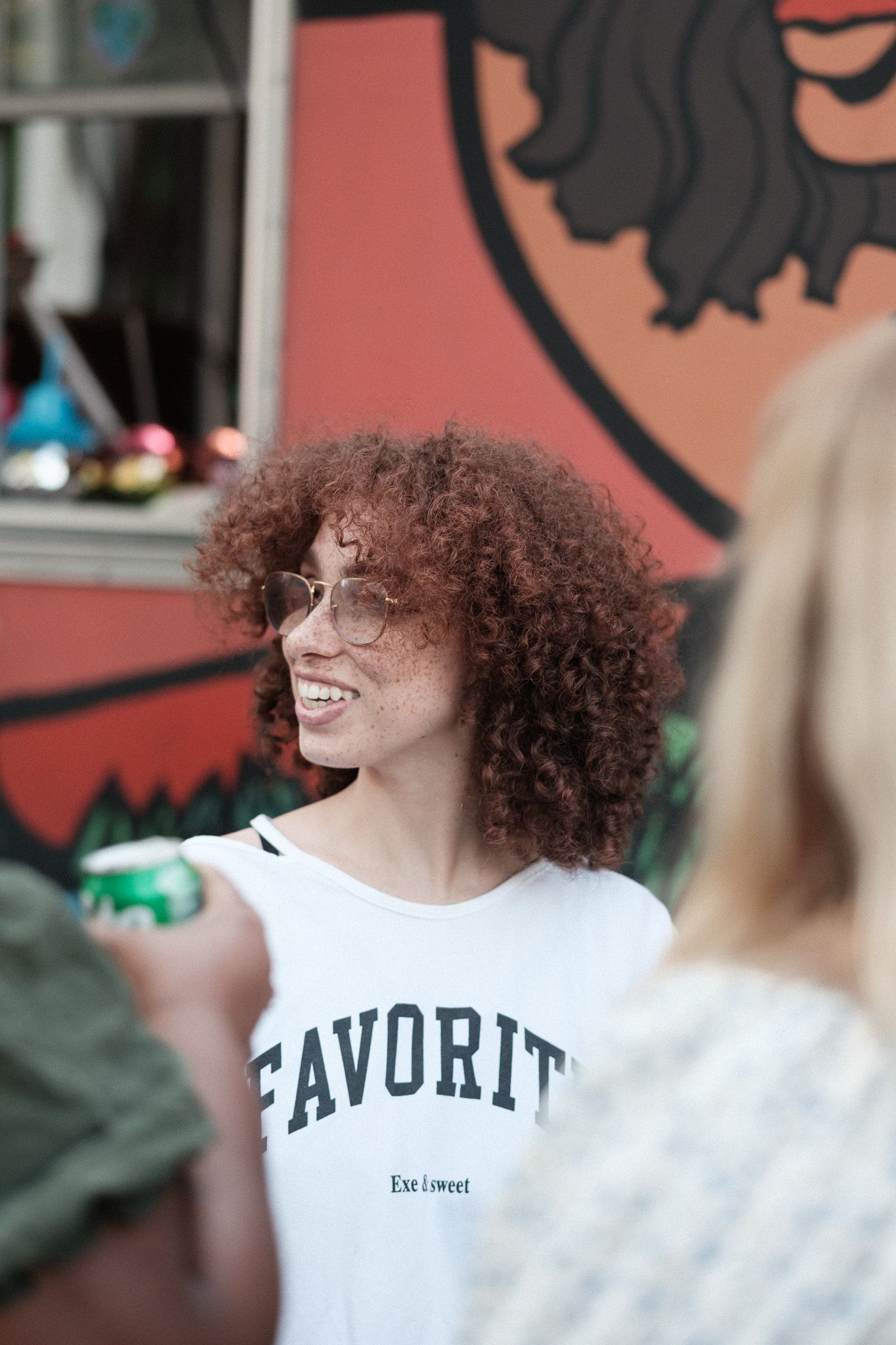 Person with curly hair wearing white t-shirt with 'FAVORITE' text smiling outdoors against pink wall.