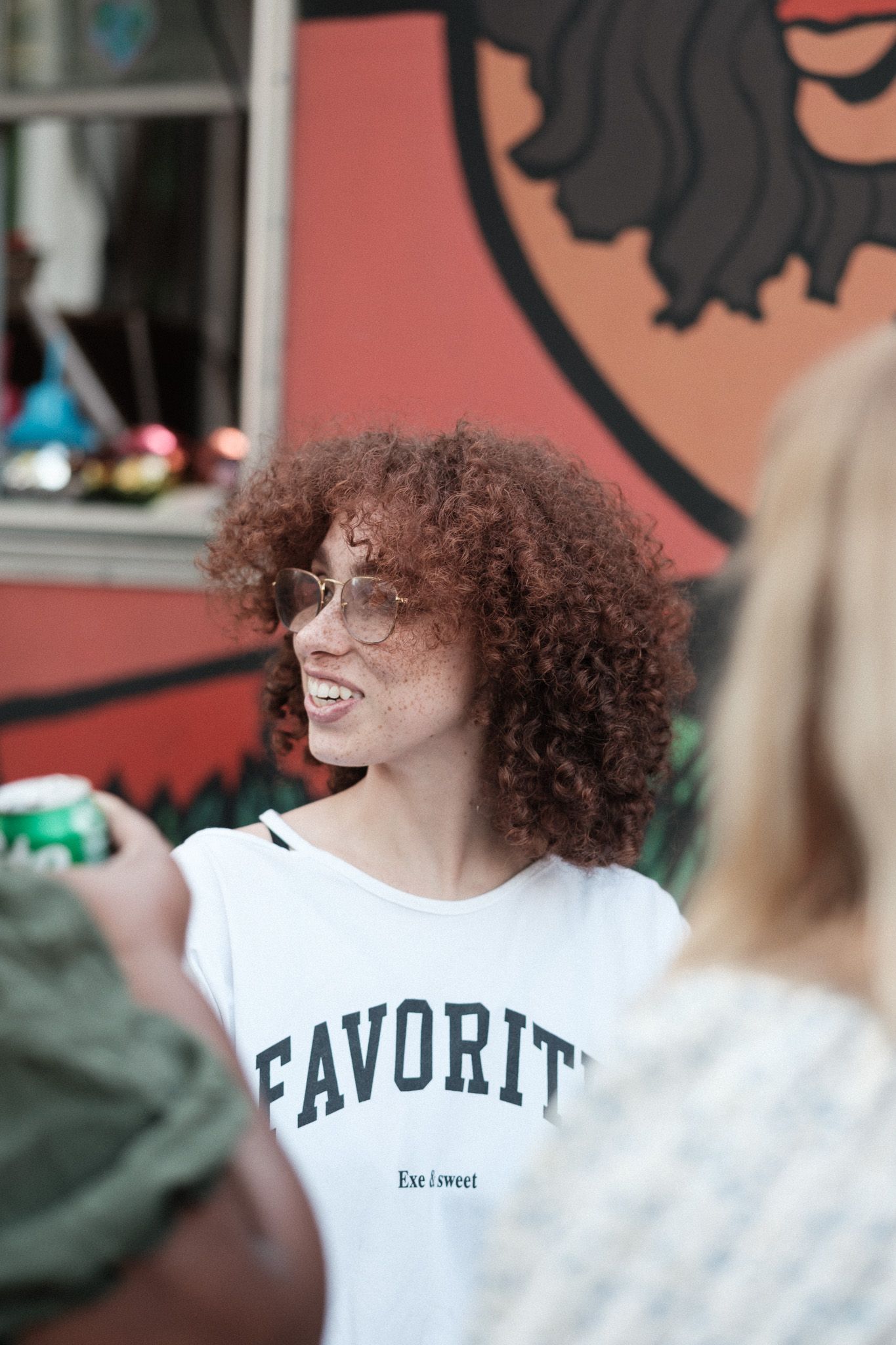 Person with curly hair wearing a white t-shirt with 'Favorite' text smiling at an outdoor event.