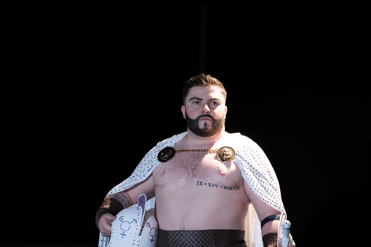 Performer in white warrior-style costume with bare chest on dark stage.