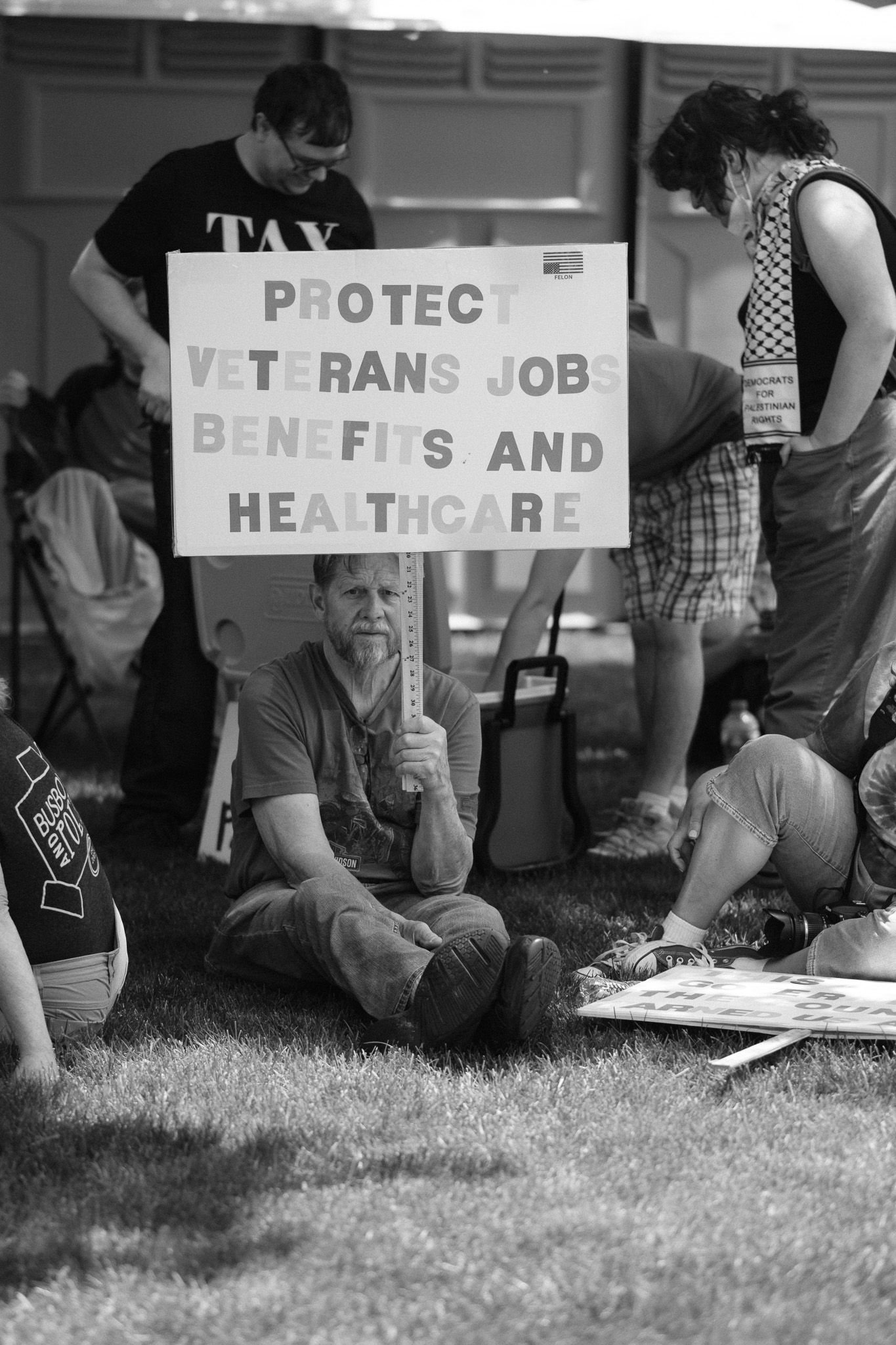 A person sits on grass holding a sign about protecting veterans' benefits and healthcare.