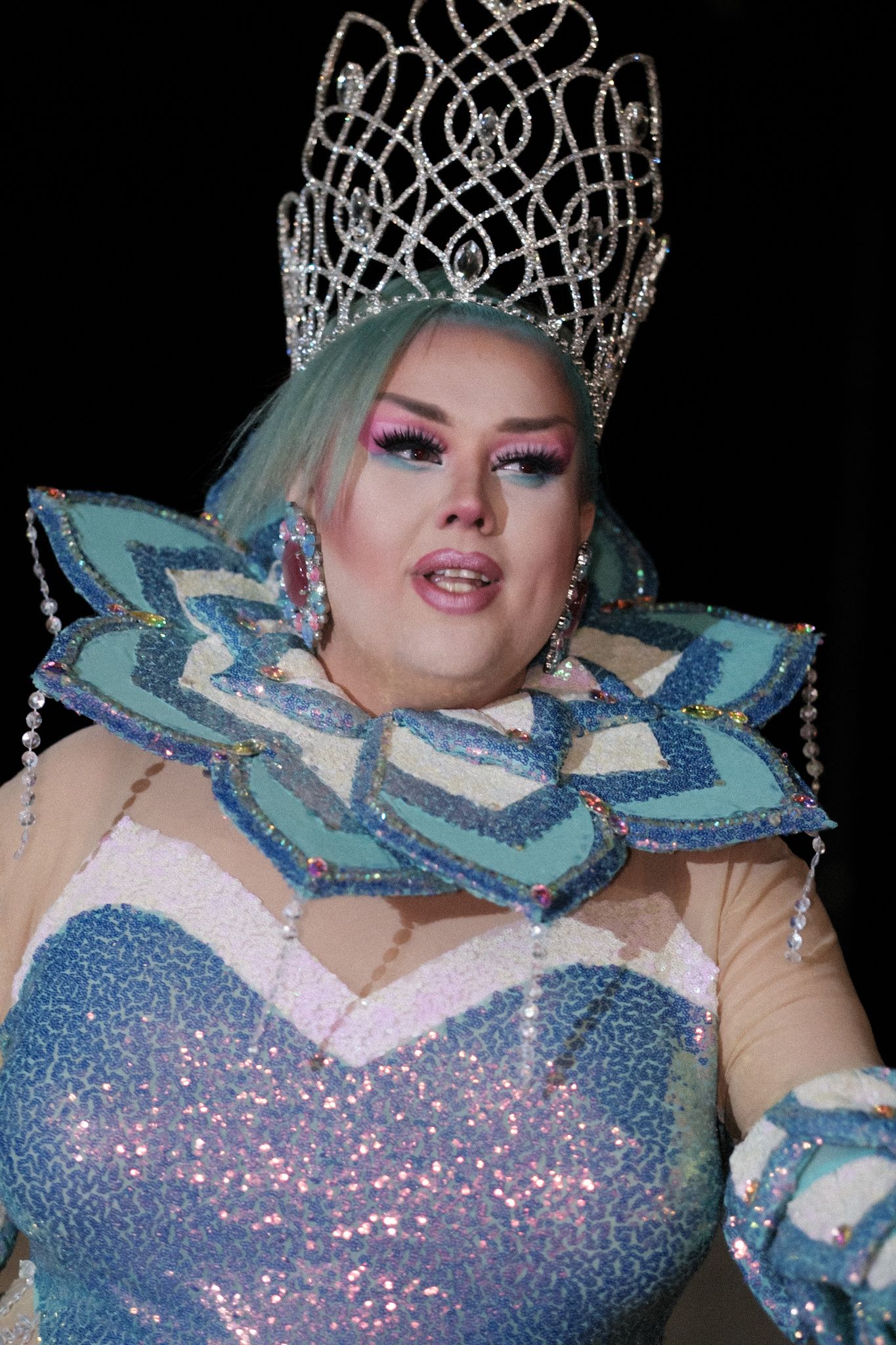 Performer wearing sparkly blue dress and ornate crown with blue and silver geometric collar piece and glittery makeup.