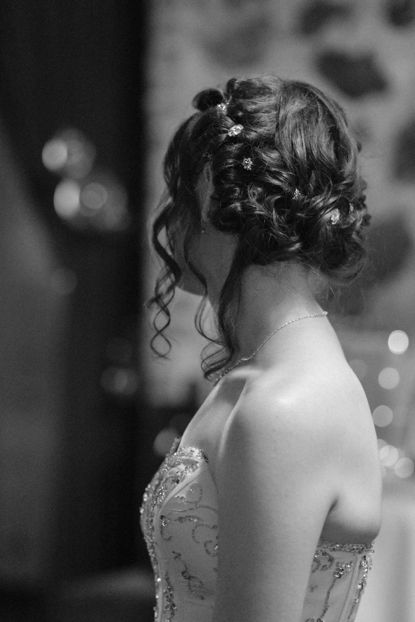 Black and white photo of an elegant updo hairstyle with flowers and curls, shown from behind in a strapless dress.