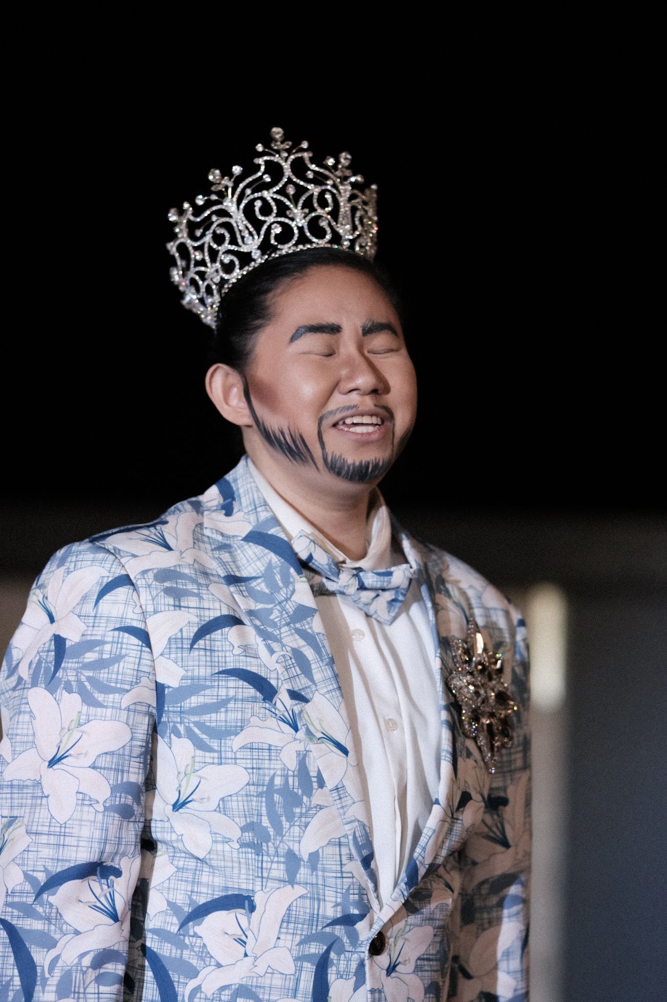 Contestant in patterned blue suit and crown smiles with eyes closed during performance.