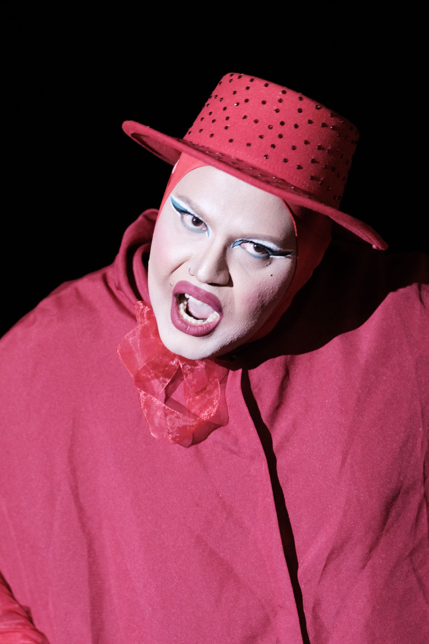 A dramatic portrait featuring bold pink attire, polka dot hat, and theatrical makeup against a black background.