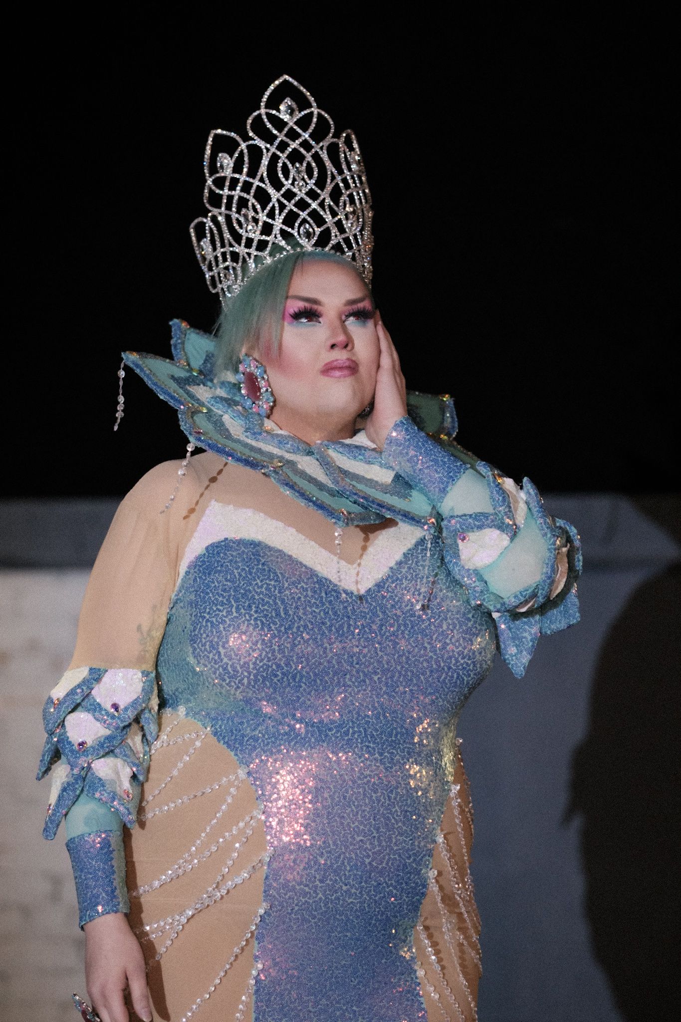 Contestant wears iridescent gown and crystal crown in dramatic stage lighting.