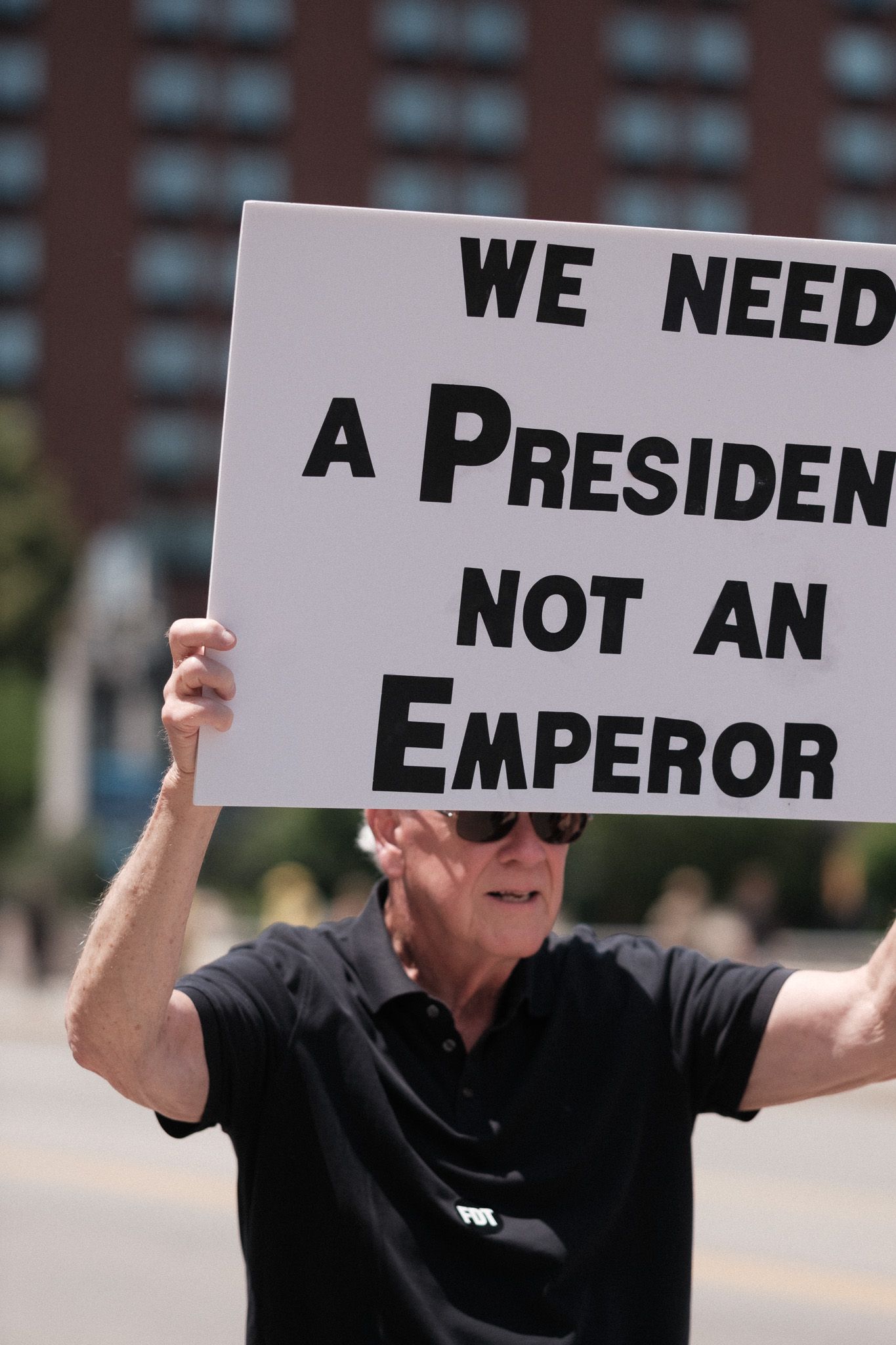 Protest sign reading 'We need a president not an emperor' held up against urban backdrop.