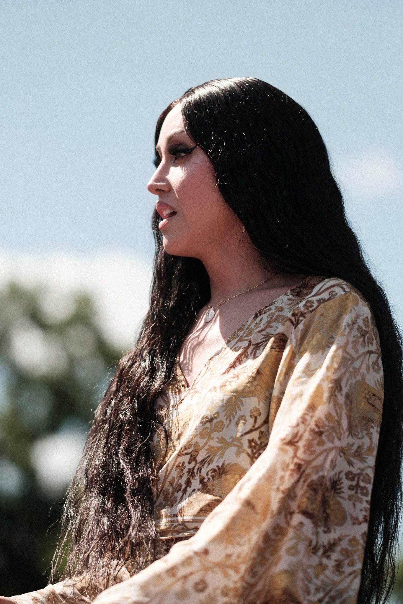 Person with very long straight dark hair wearing an off-white patterned dress photographed in profile against a blue sky.
