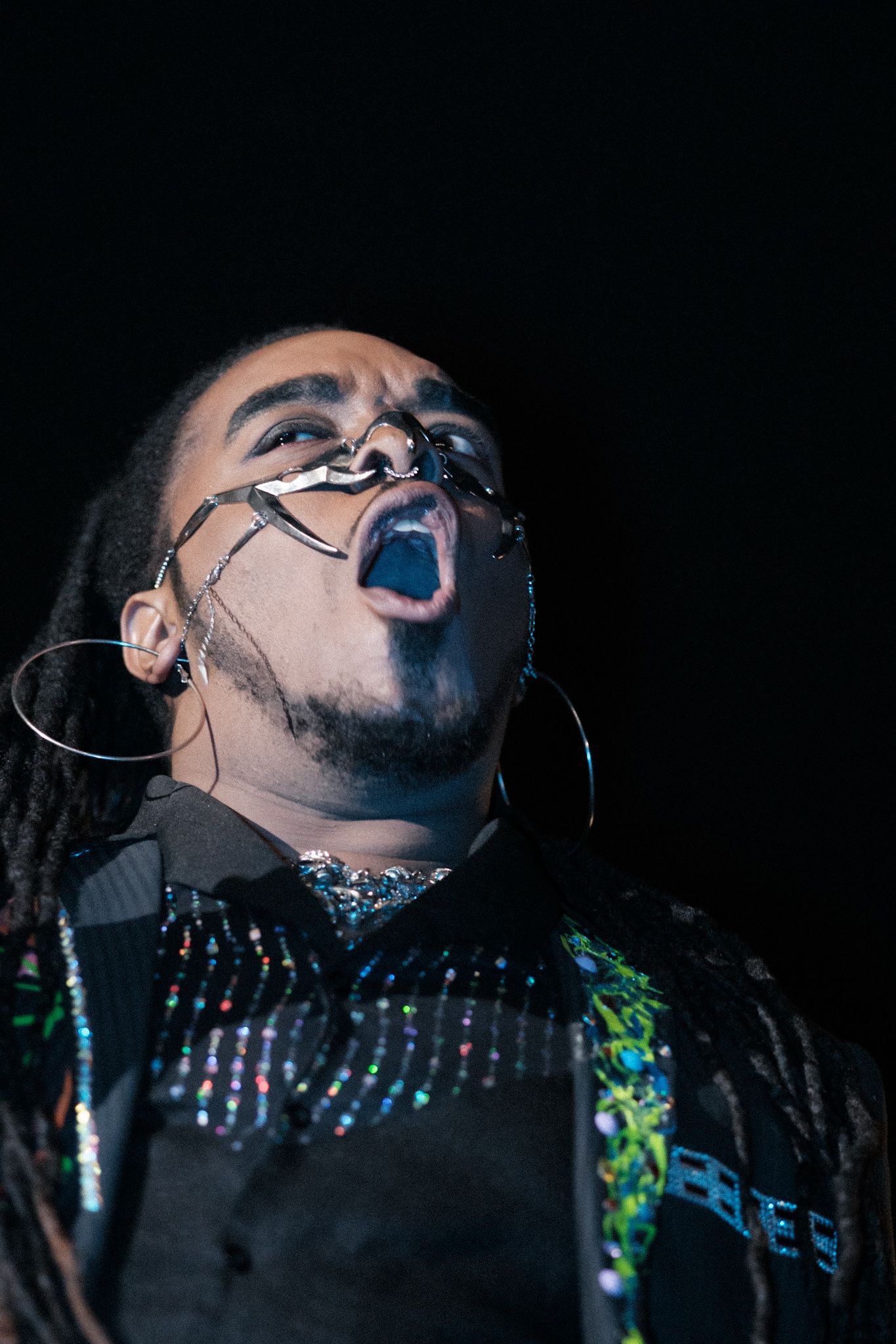 Performer in dark clothing and face paint singing passionately against black background.
