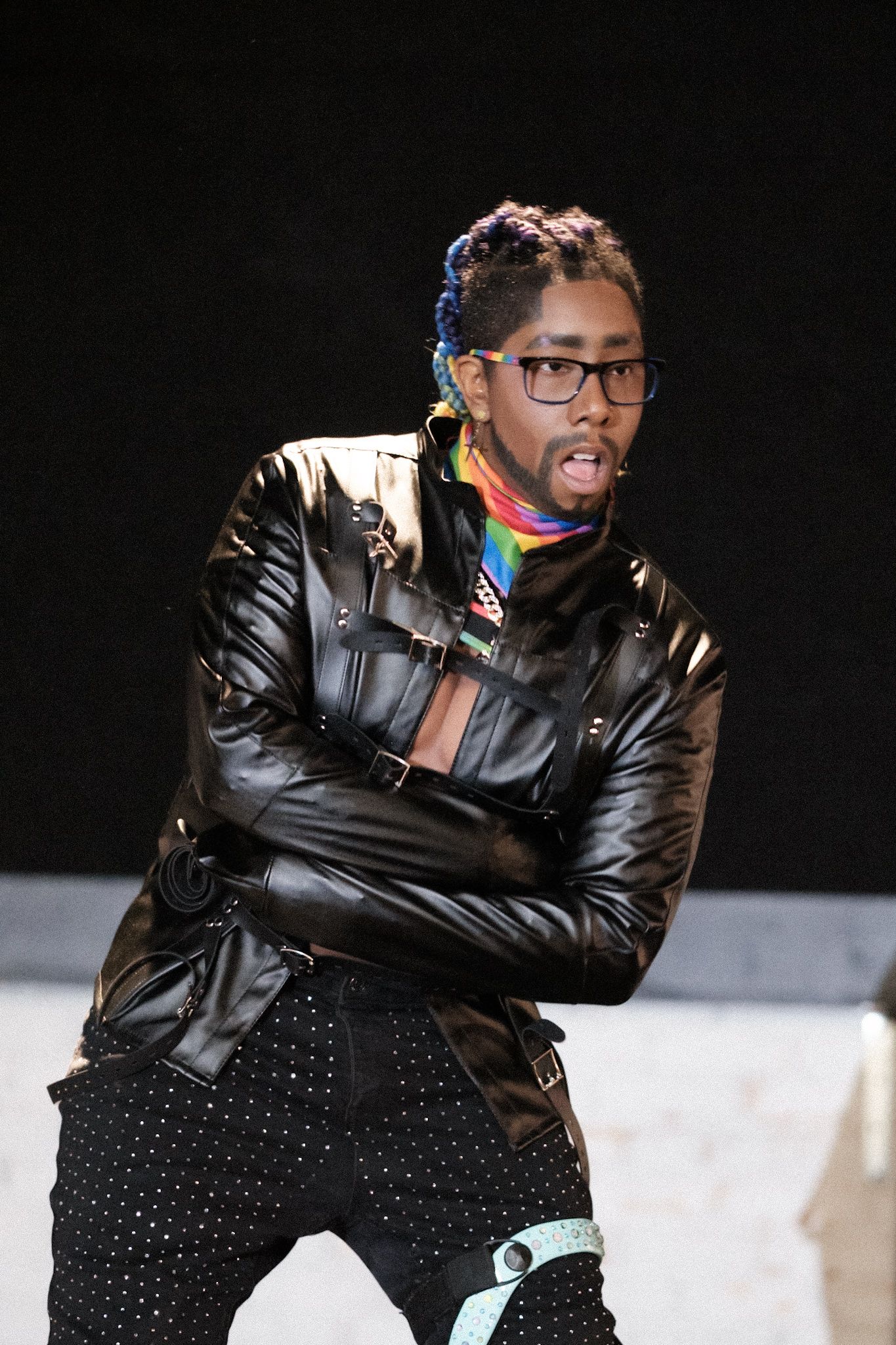 A performer in a black leather jacket and polka dot pants poses on stage.