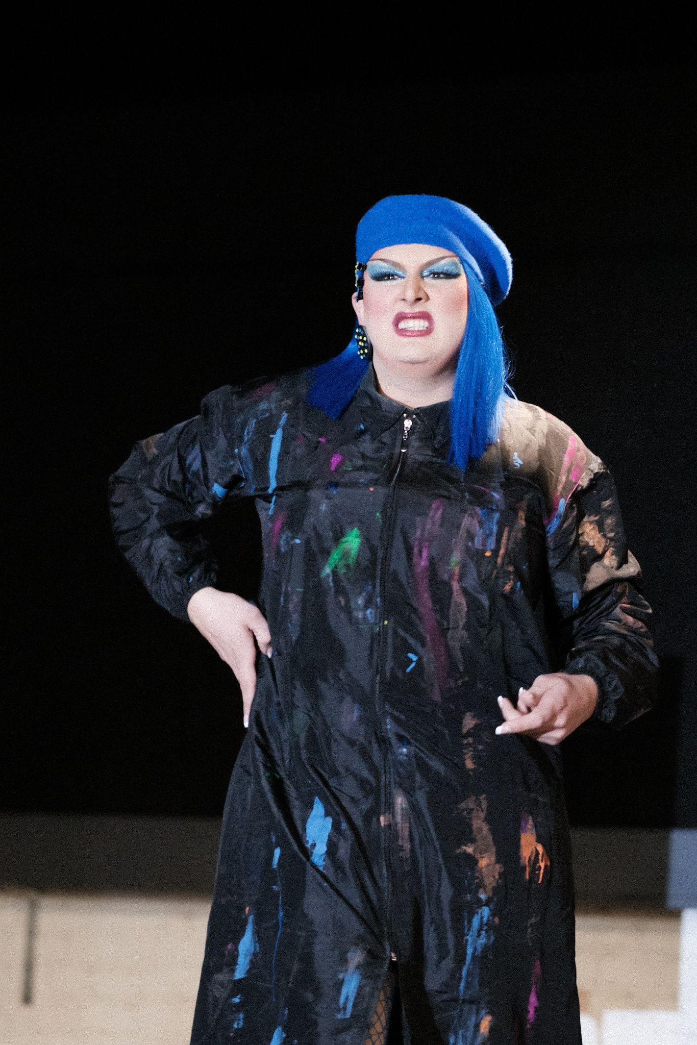 Performer in a vibrant blue beret and black paint-splattered coat strikes a dramatic pose on stage.