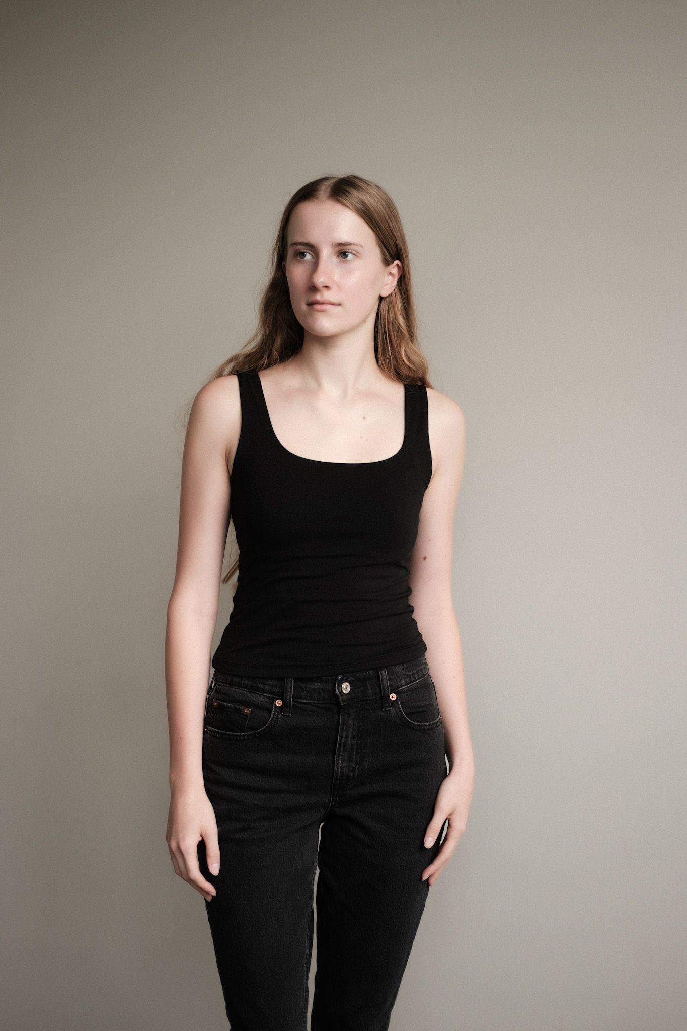 A person wearing a black tank top and dark jeans stands against a neutral wall in a minimalist portrait.