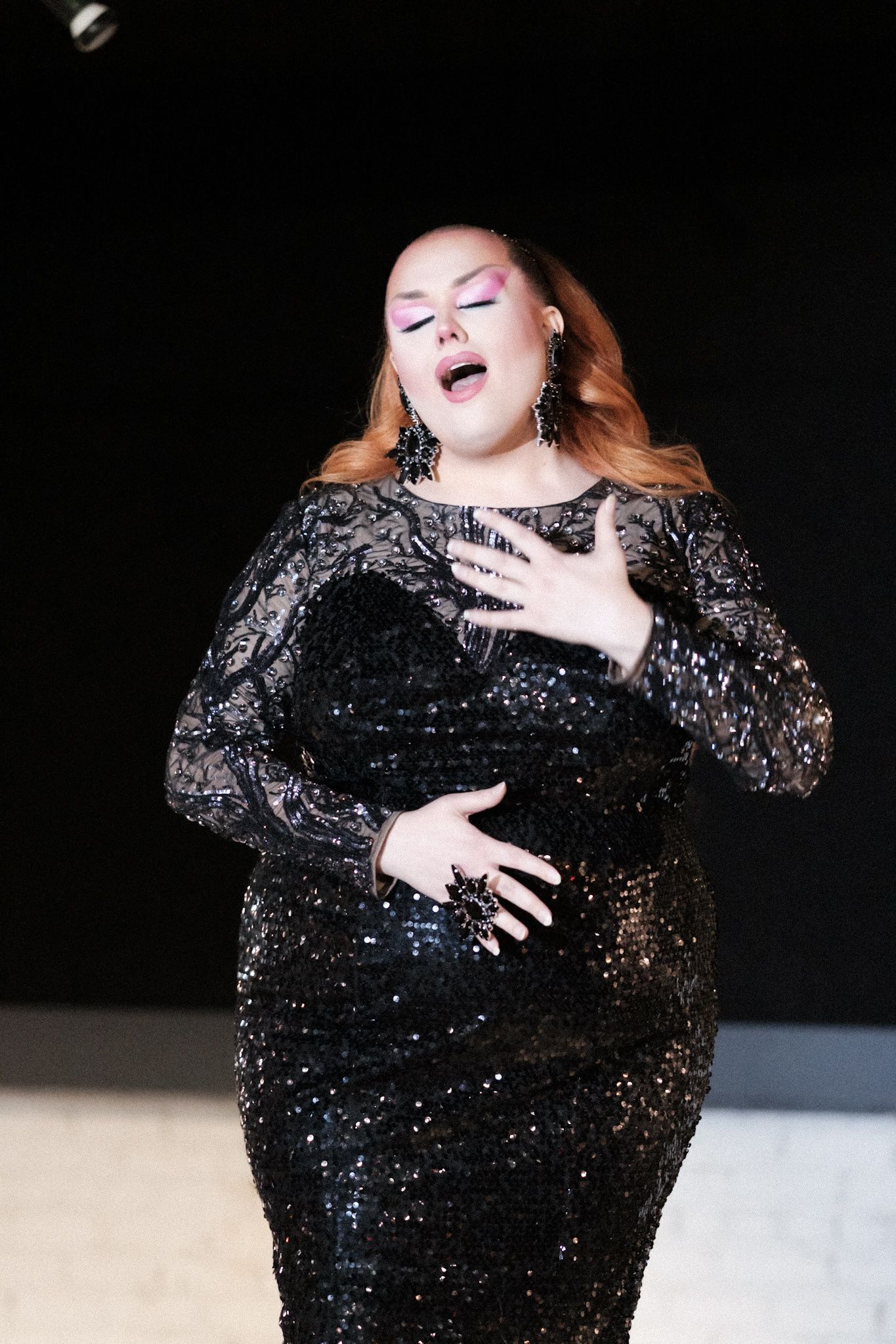 A performer in a sparkling black sequined gown singing passionately on stage.