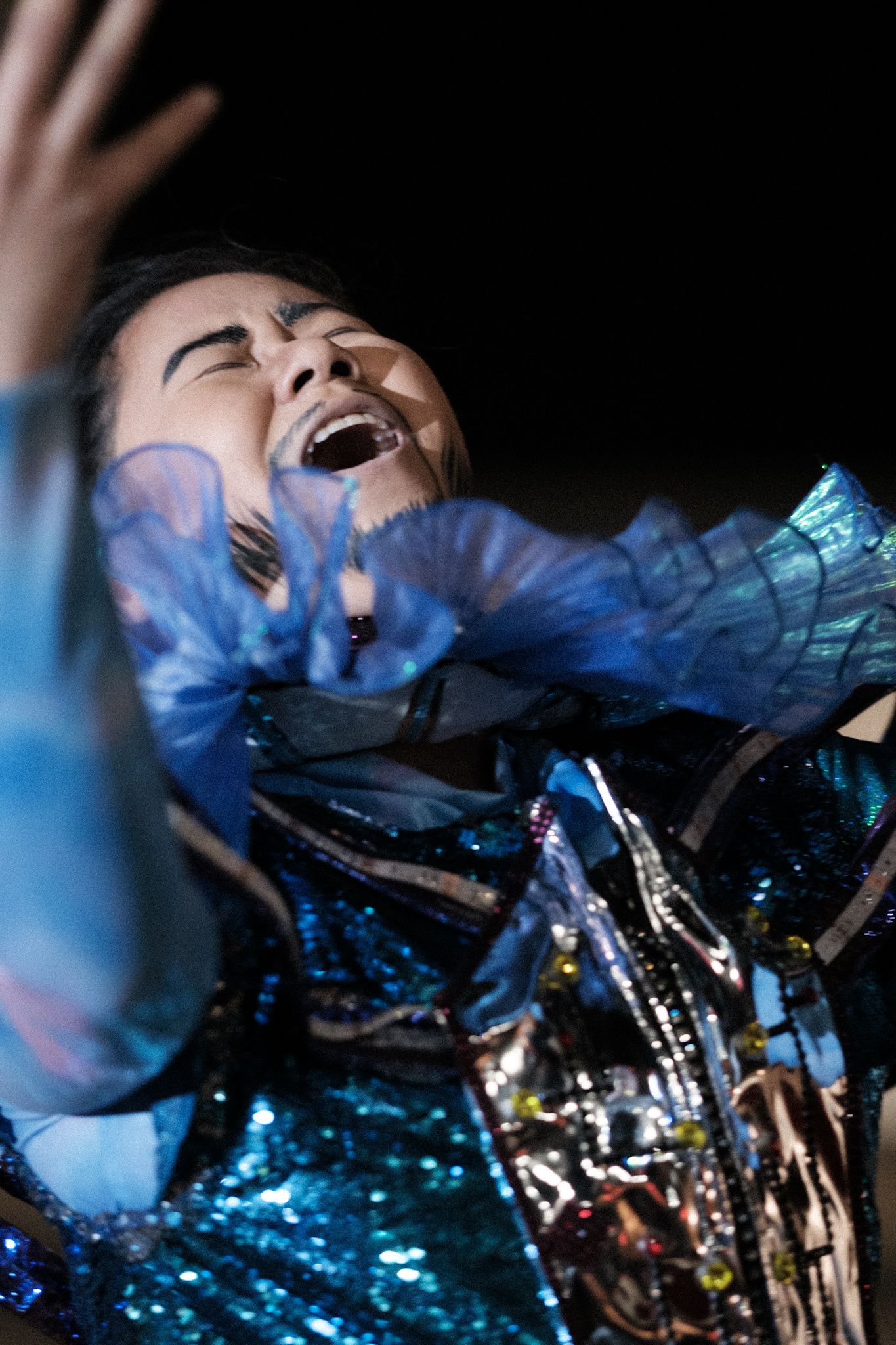 Performer in blue and silver attire expresses joy with upward gaze and raised hand.