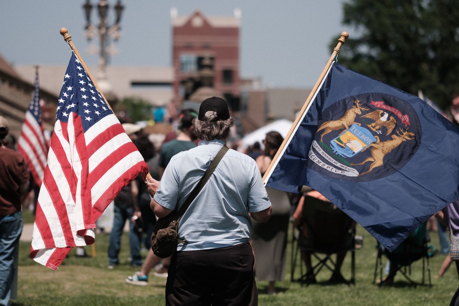 Person holding American flag and Michigan state flag at outdoor rally gathering with building in background.