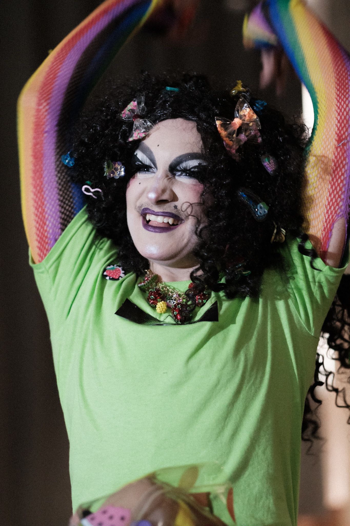 Performer in green shirt and curly black hair with colorful decorations smiling during theatrical performance.