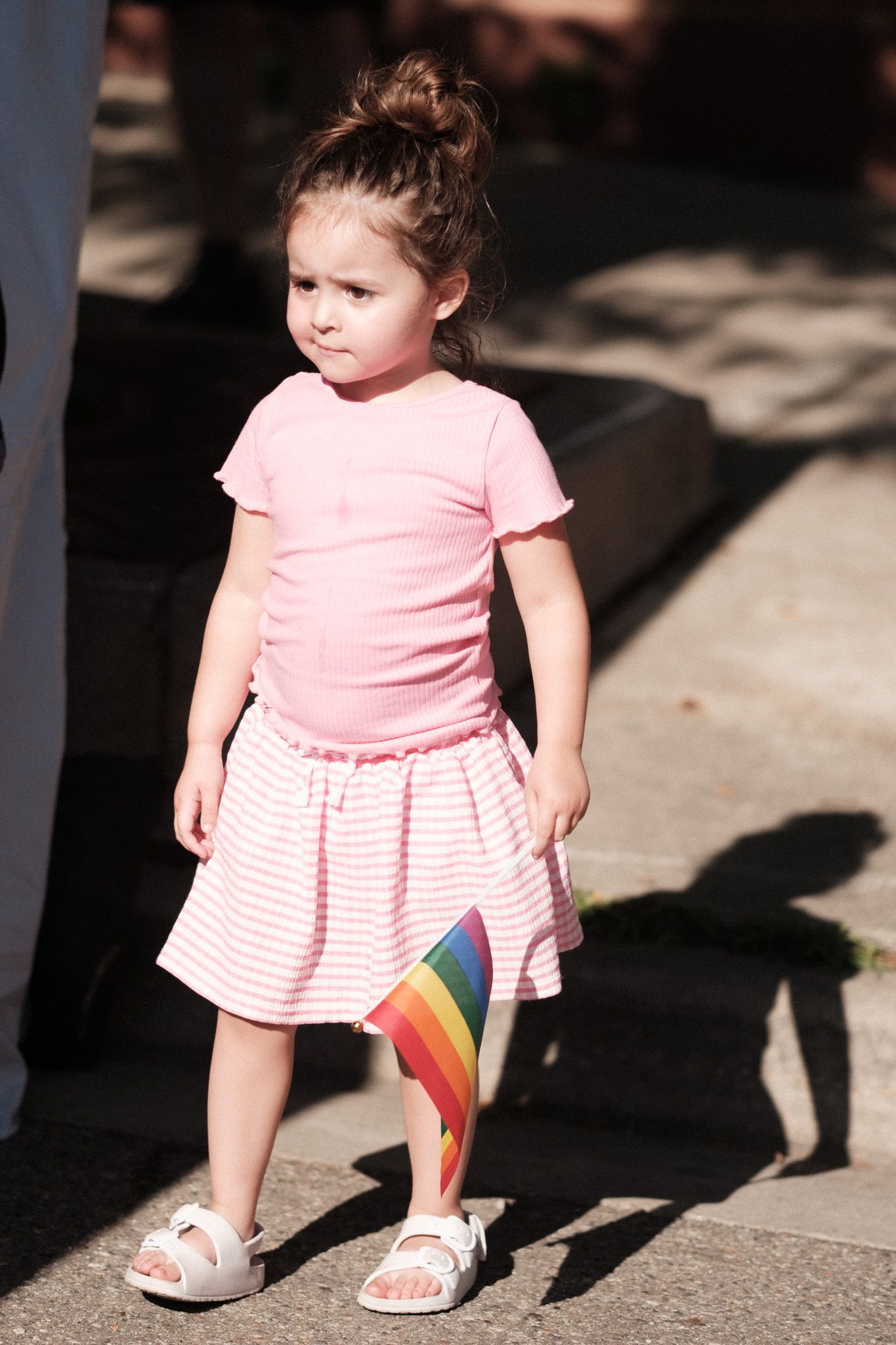 A small child in a pink dress and flip flops stands on a sunny sidewalk holding a rainbow flag.