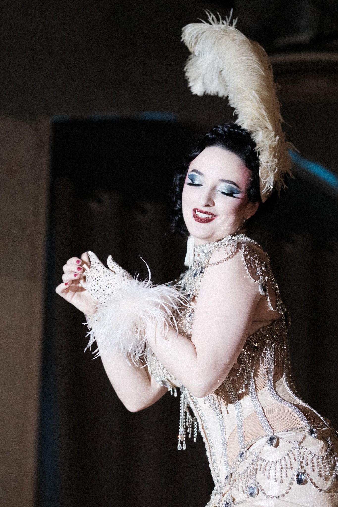 Burlesque performer in a glamorous white feathered and rhinestone costume strikes a dramatic pose on stage.