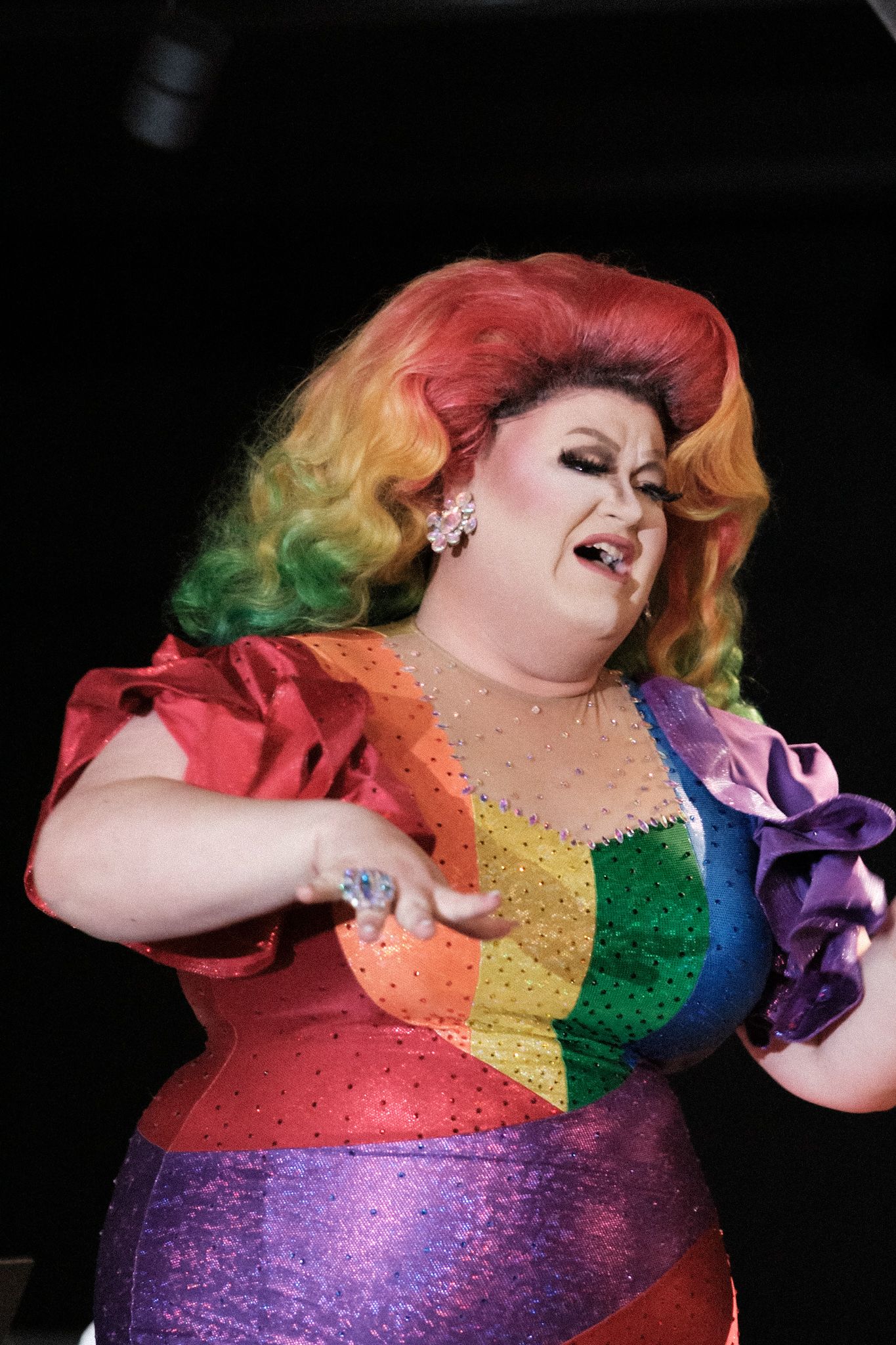 A drag performer in a rainbow-colored sequin costume performs dynamically on a dark stage.