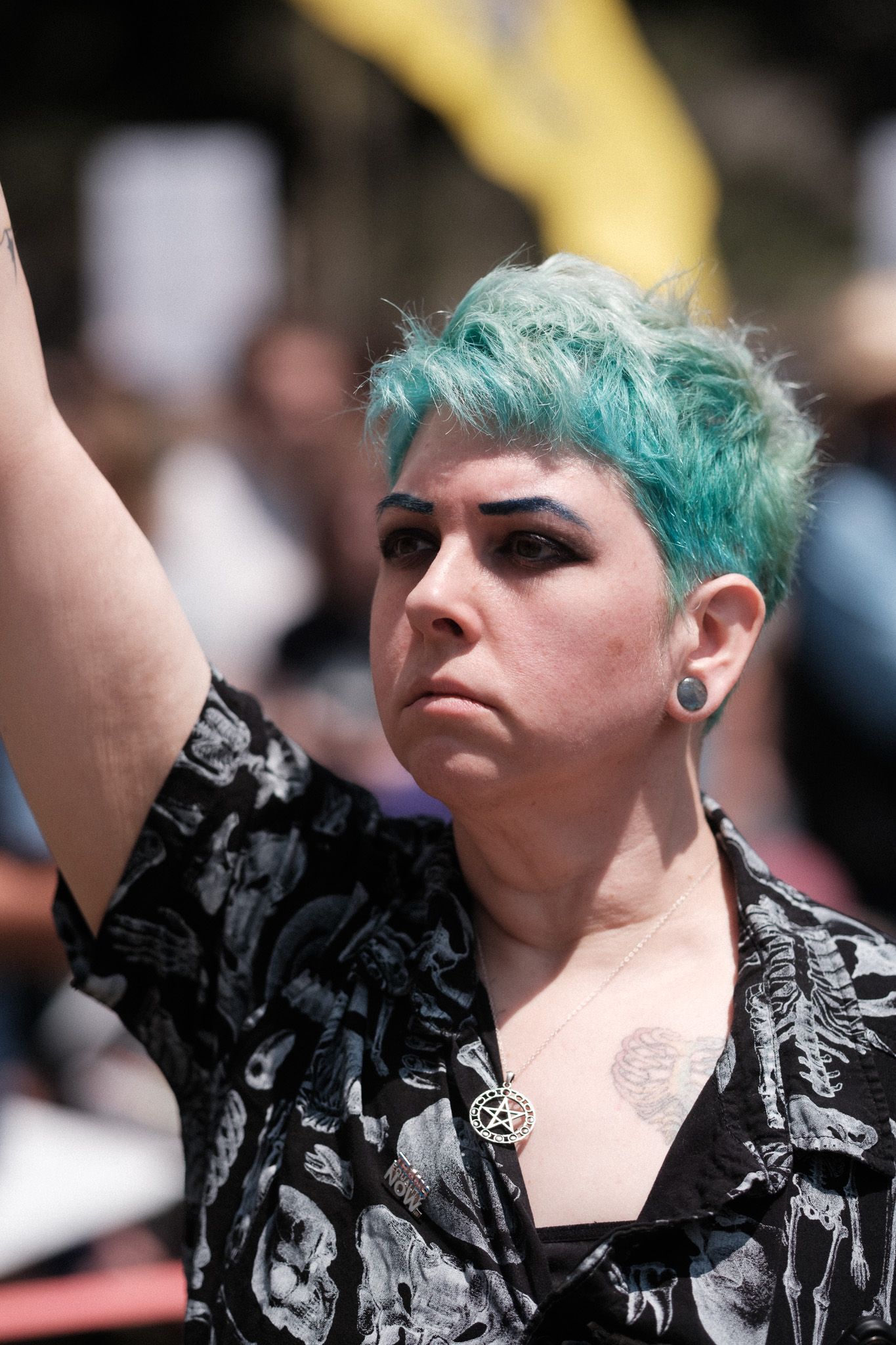 A person with teal hair and piercings raises their fist during a protest gathering.