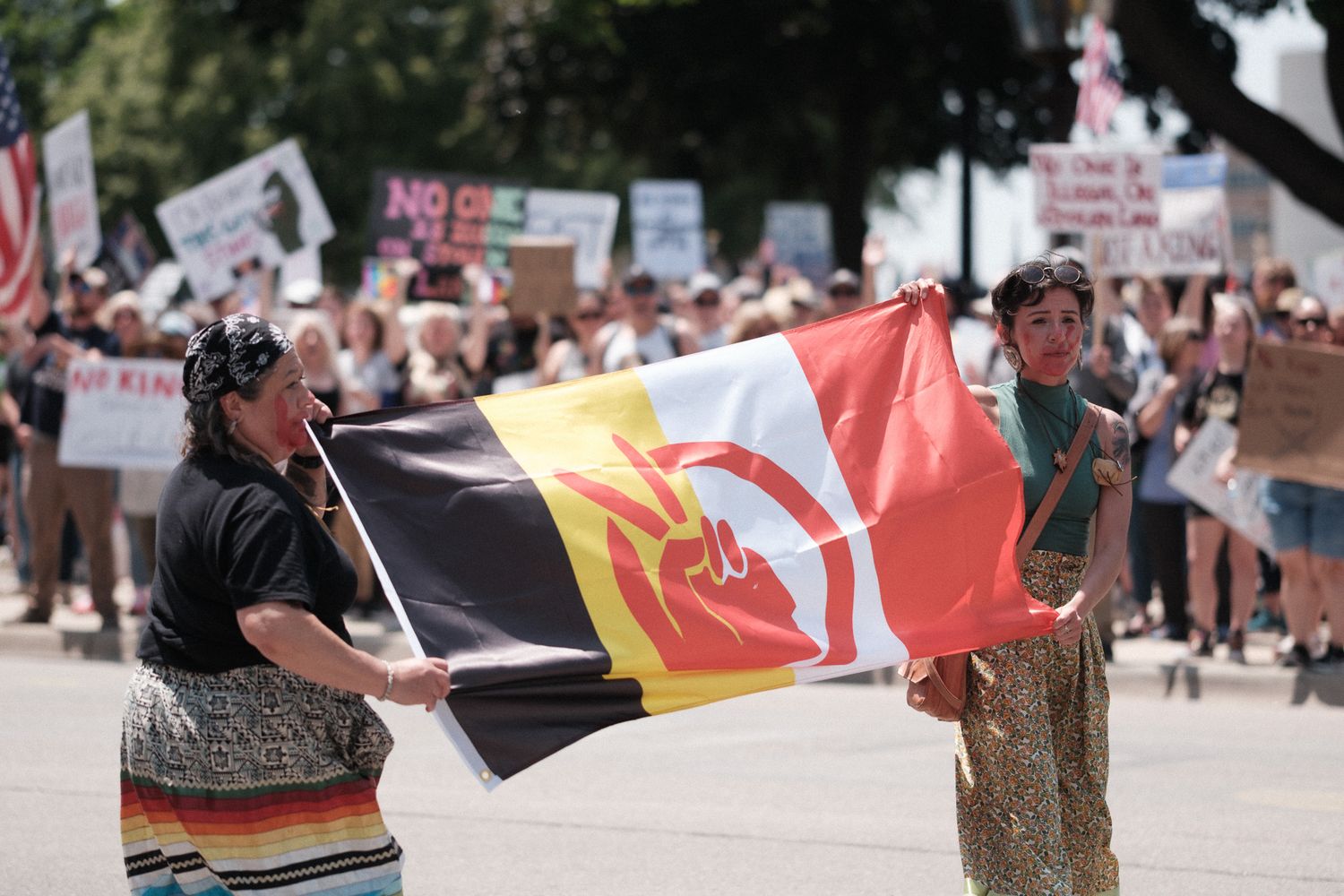 Protesters hold an American Indian Movement flag during an outdoor demonstration on a sunny day.