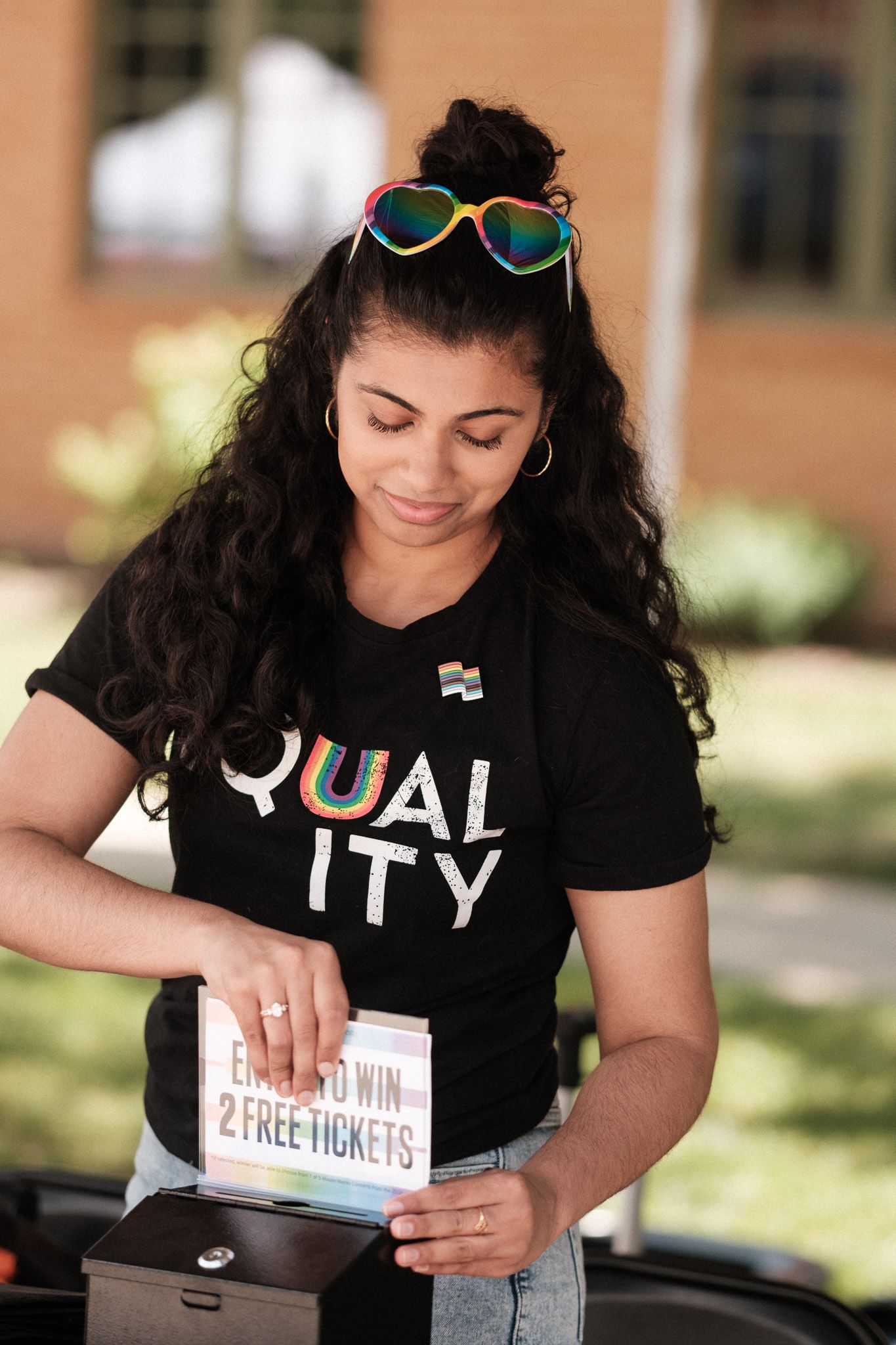 Person wearing a Quality t-shirt and rainbow sunglasses looks down at tickets while standing outdoors.
