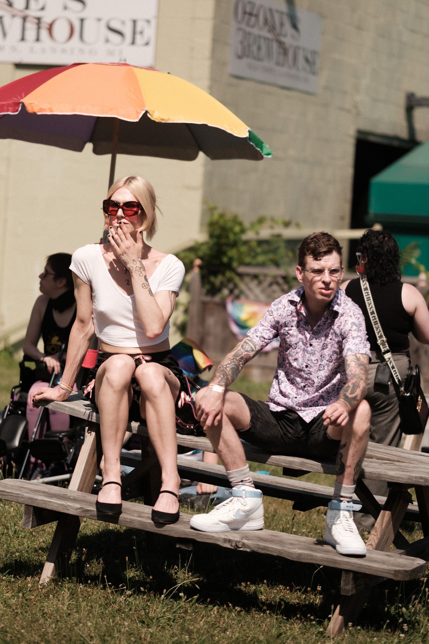Two festival attendees relax on a wooden picnic table under a colorful umbrella on a sunny day.