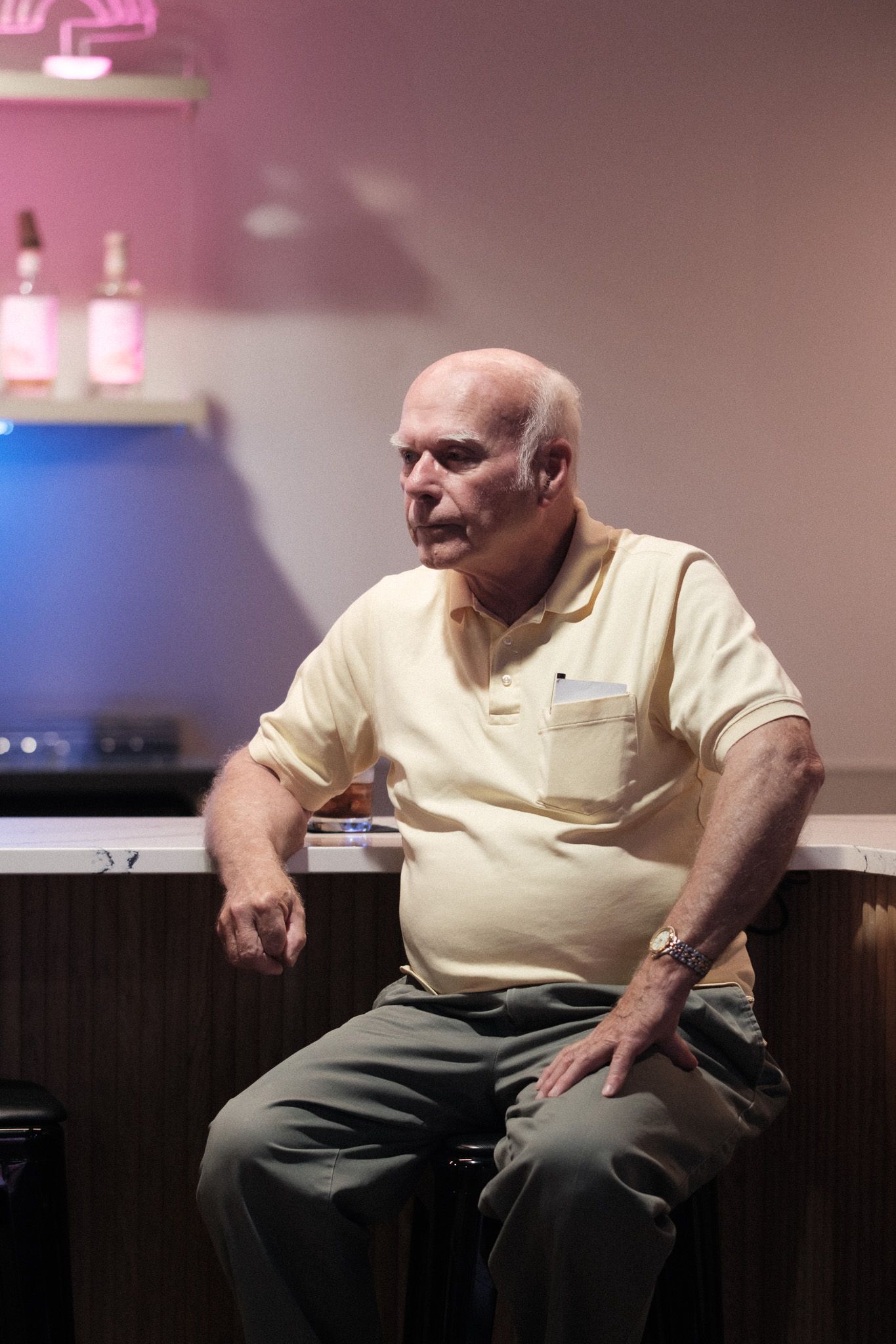 Someone wearing a yellow shirt and grey pants sits on a dark bench in a room with pink-tinted lighting.