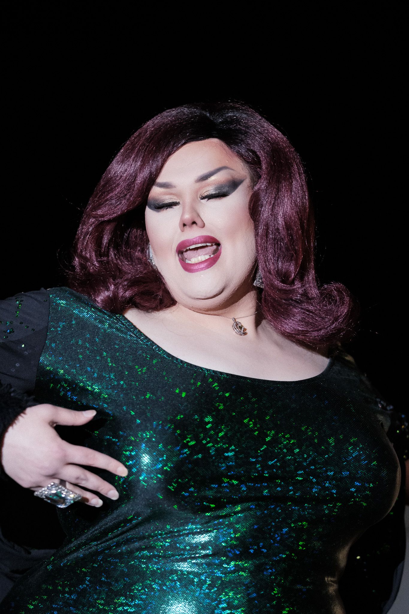 A performer wearing a sparkly emerald green sequined gown and dark wig poses dramatically against a dark background.