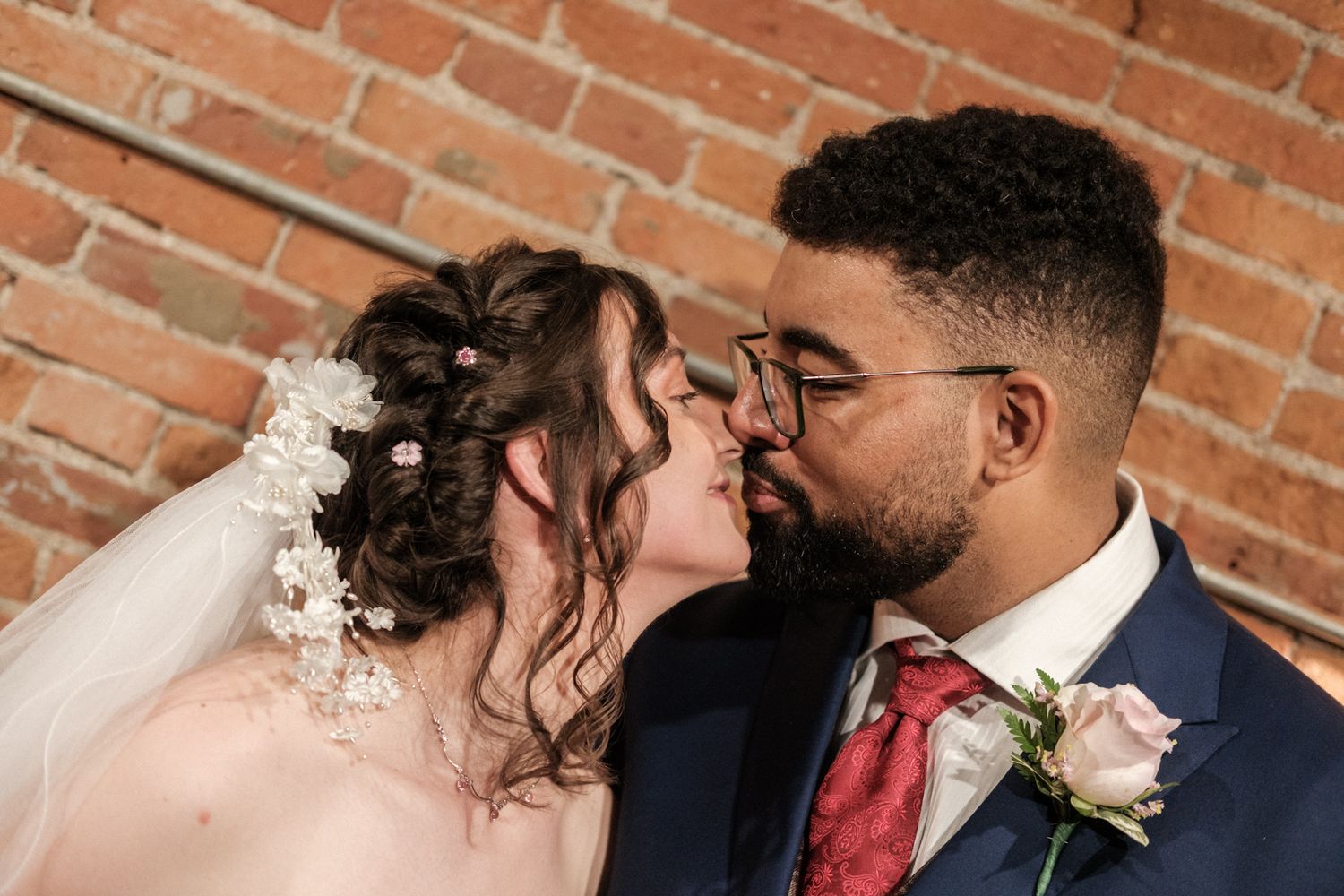 Close up portrait of newlyweds sharing intimate moment in front of brick wall, bride wearing veil and groom in navy suit.