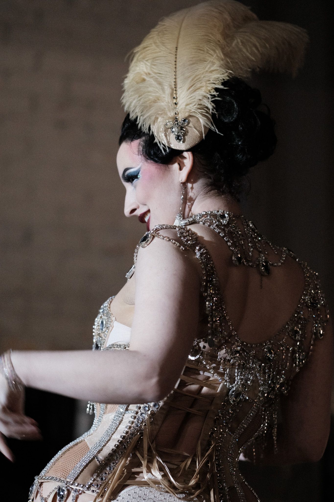 A performer in an ornate vintage burlesque costume with feathers and crystals poses dramatically in low light.