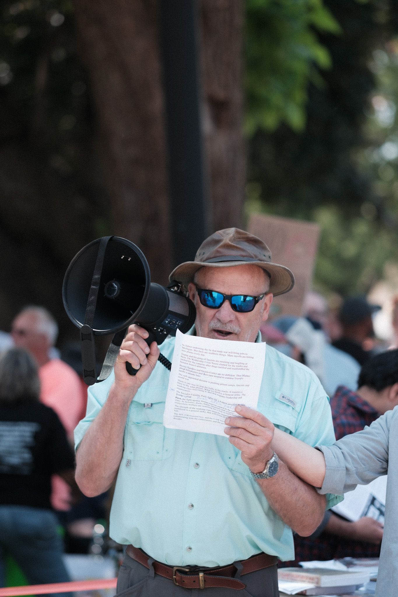 A protester in a light blue shirt holds papers while speaking through a megaphone.