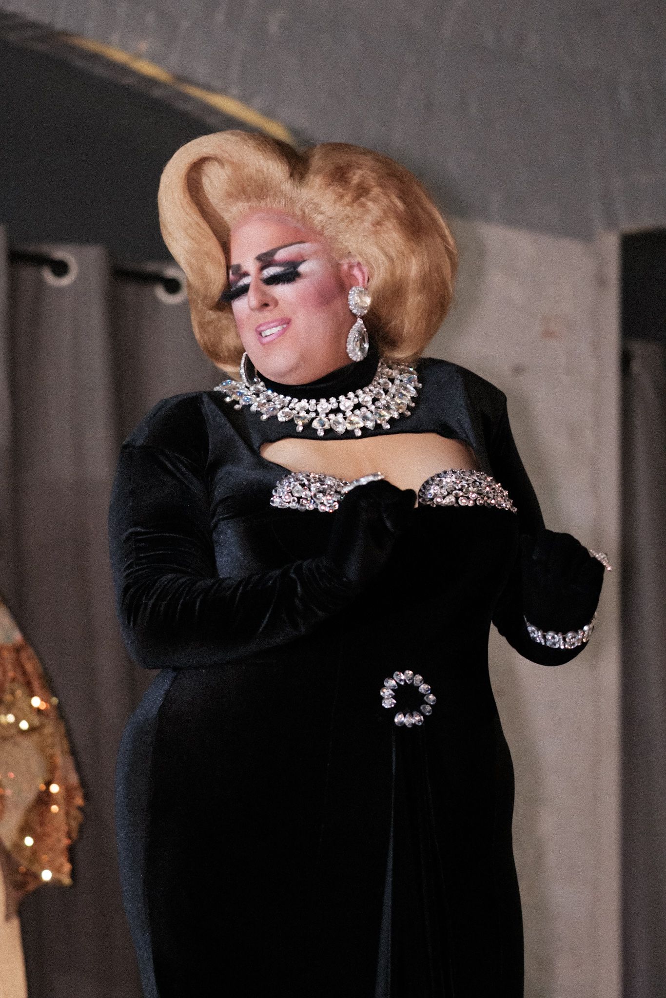 Glamorous performer in black velvet gown with ornate crystal necklace and dramatic blonde updo.