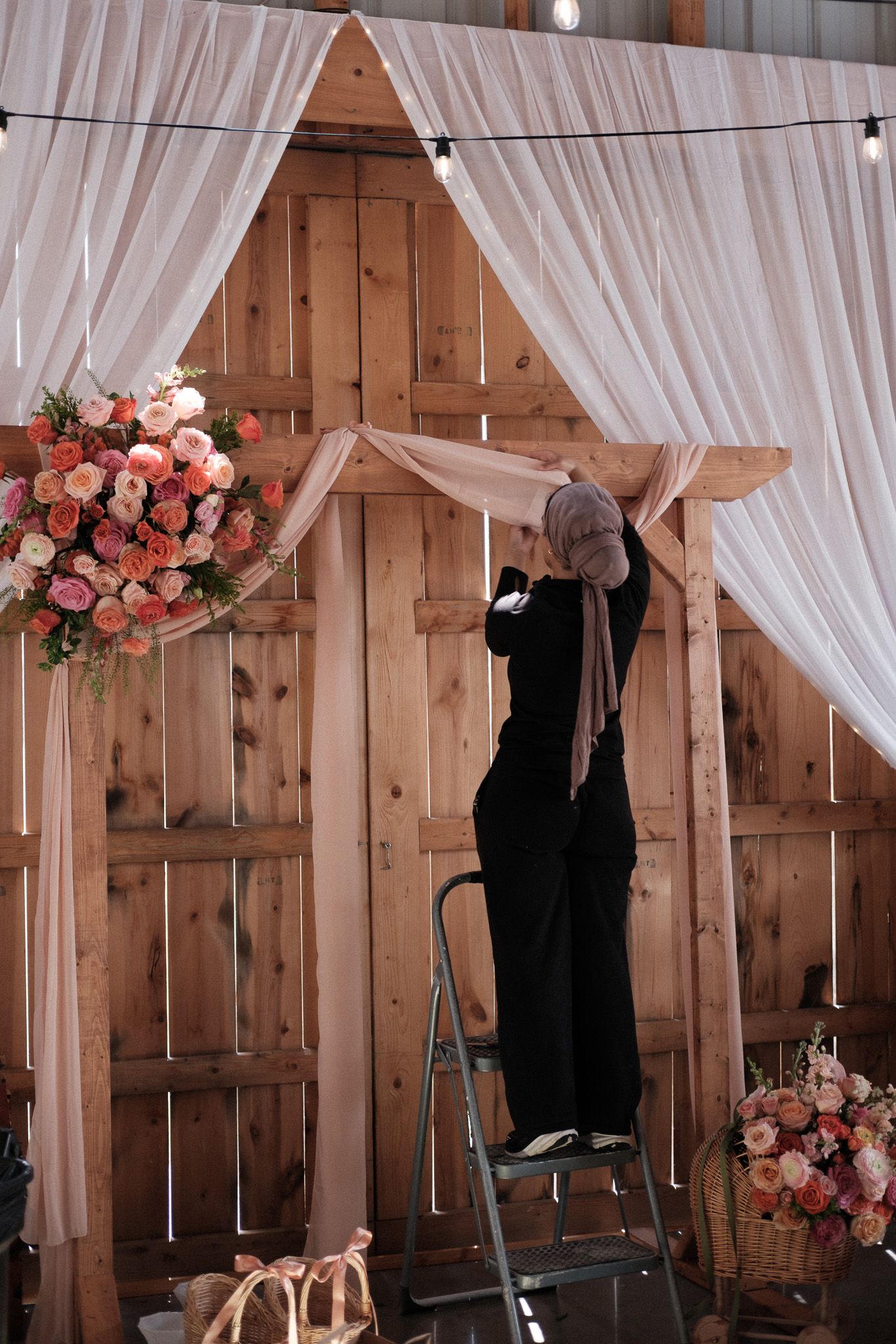 Event planner on ladder decorates rustic barn venue with white drapes and pink floral arrangements.