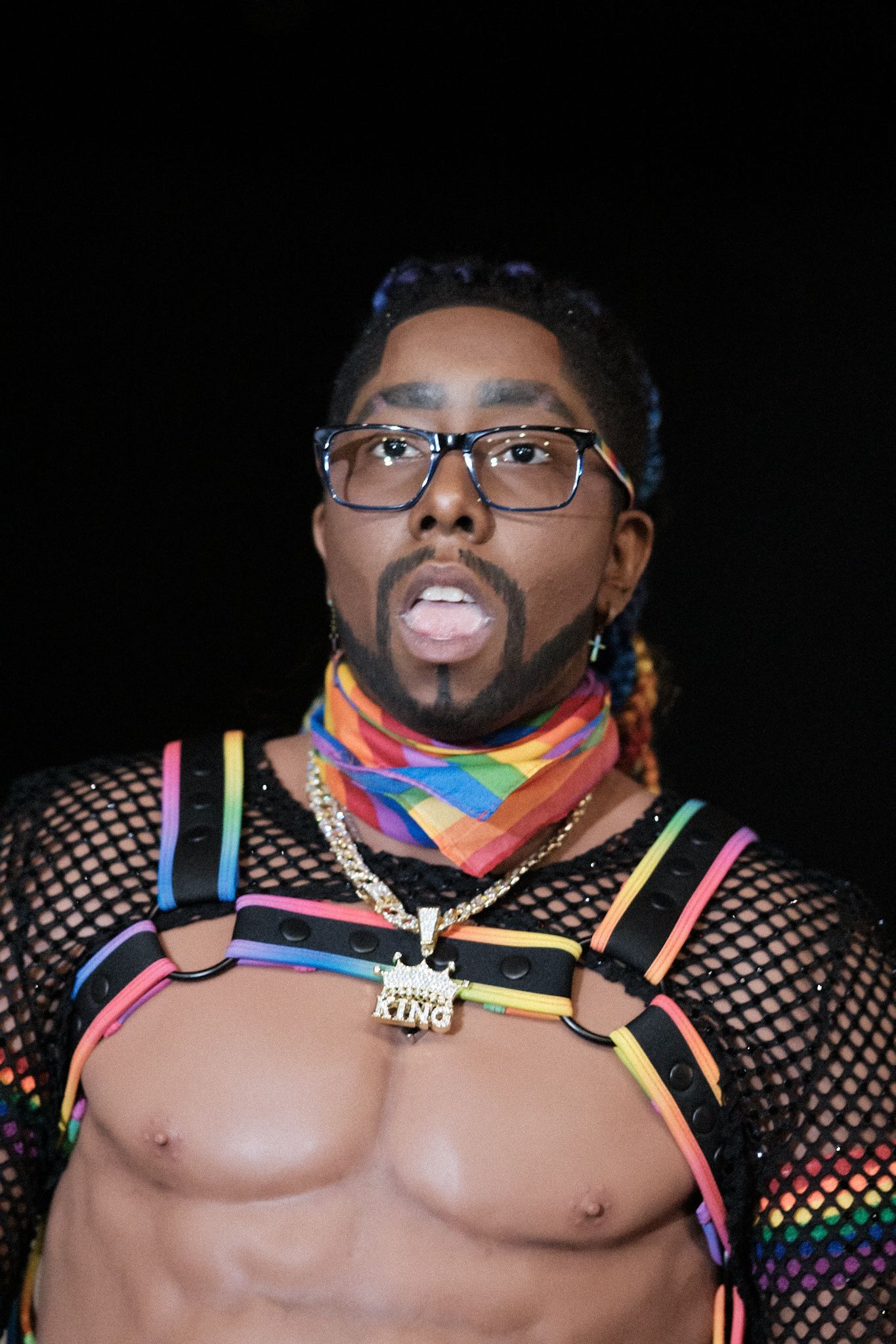 Performer in rainbow mesh top with pride scarf and glasses against dark background.
