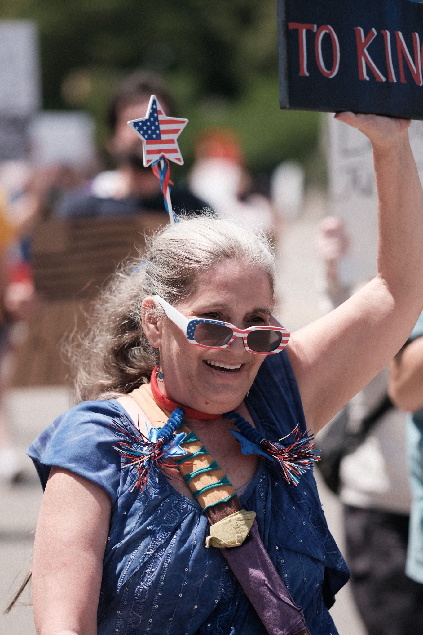 Demonstrators smile and wave flags while holding protest signs during an outdoor rally.