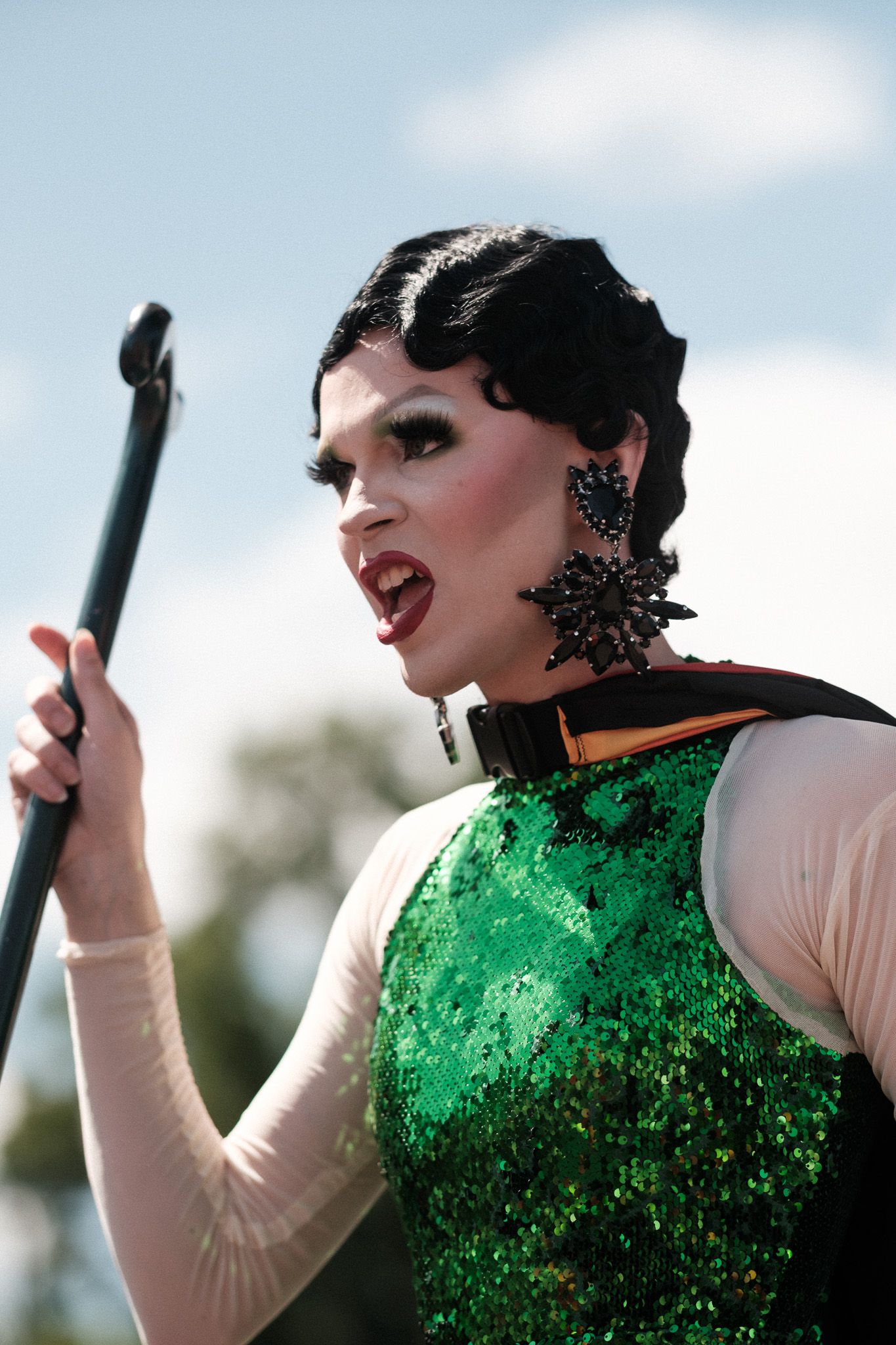 Performer in a sparkly green and white costume holds a microphone stand during an outdoor show.
