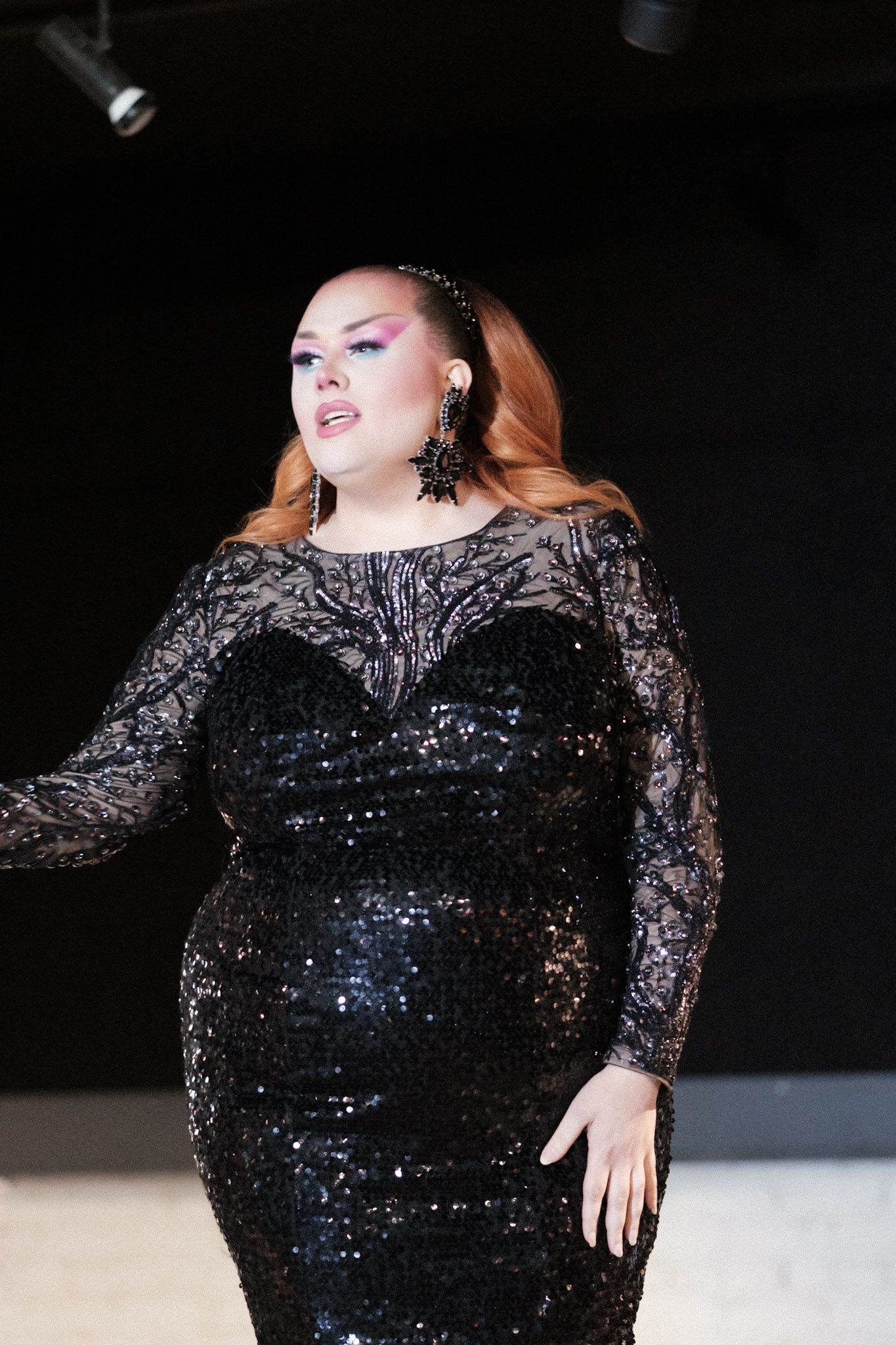 Elegant performer in black sequined gown with lace sleeves poses dramatically on stage.