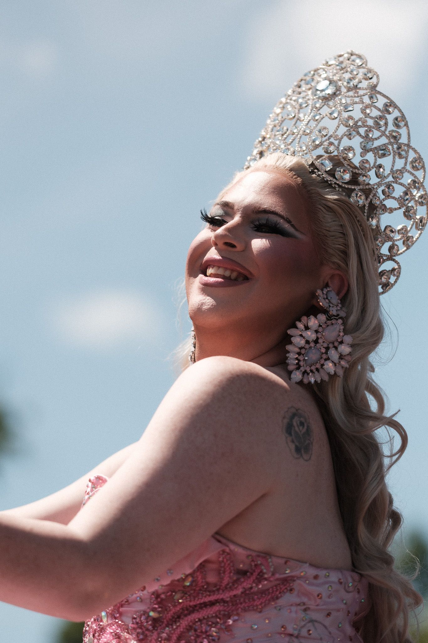 A performer wearing an ornate crystal crown and pink dress smiles during a sunny outdoor celebration.