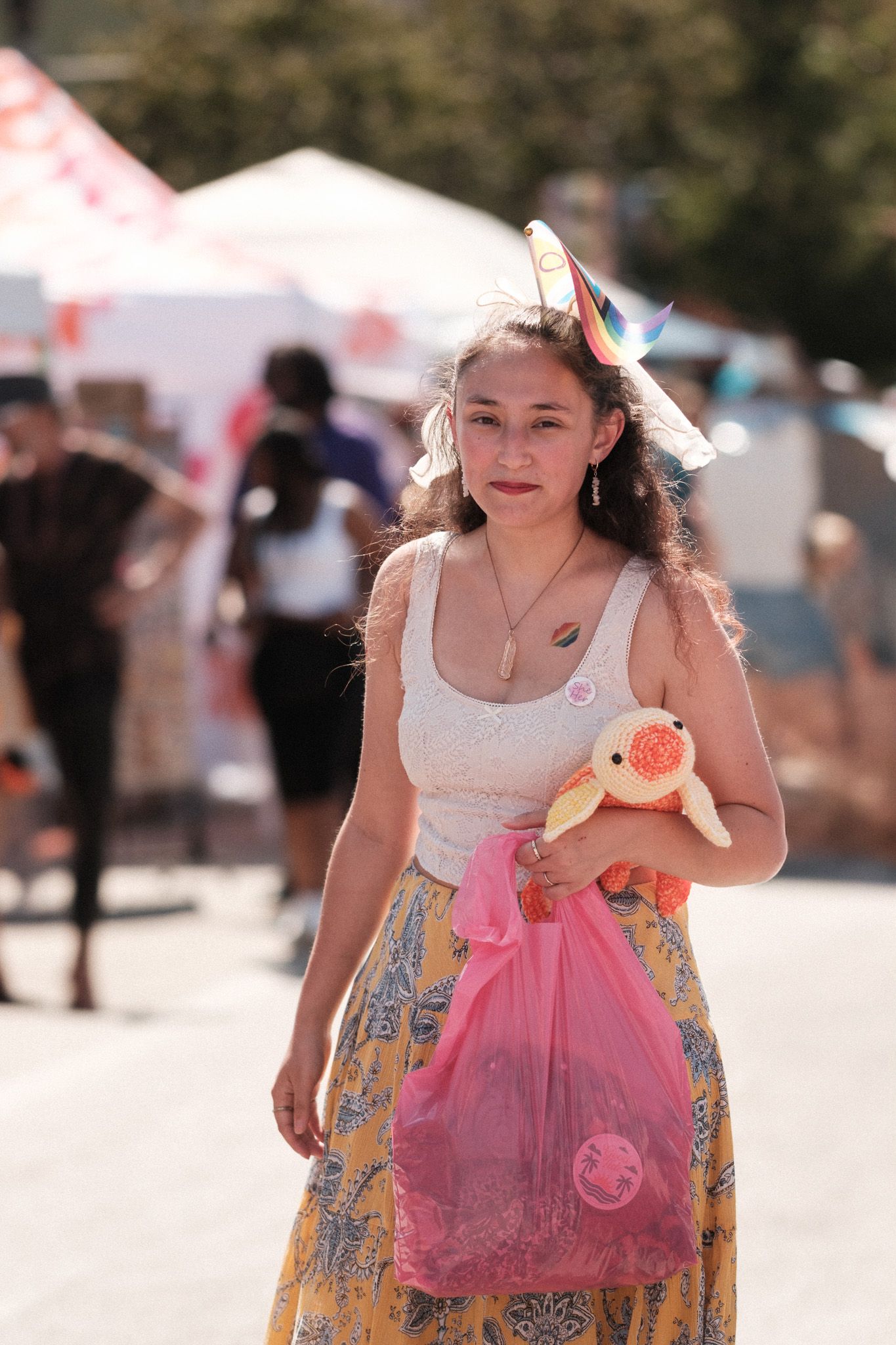 Young festival goer in a floral skirt and white top carrying a pink shopping bag at an outdoor event.