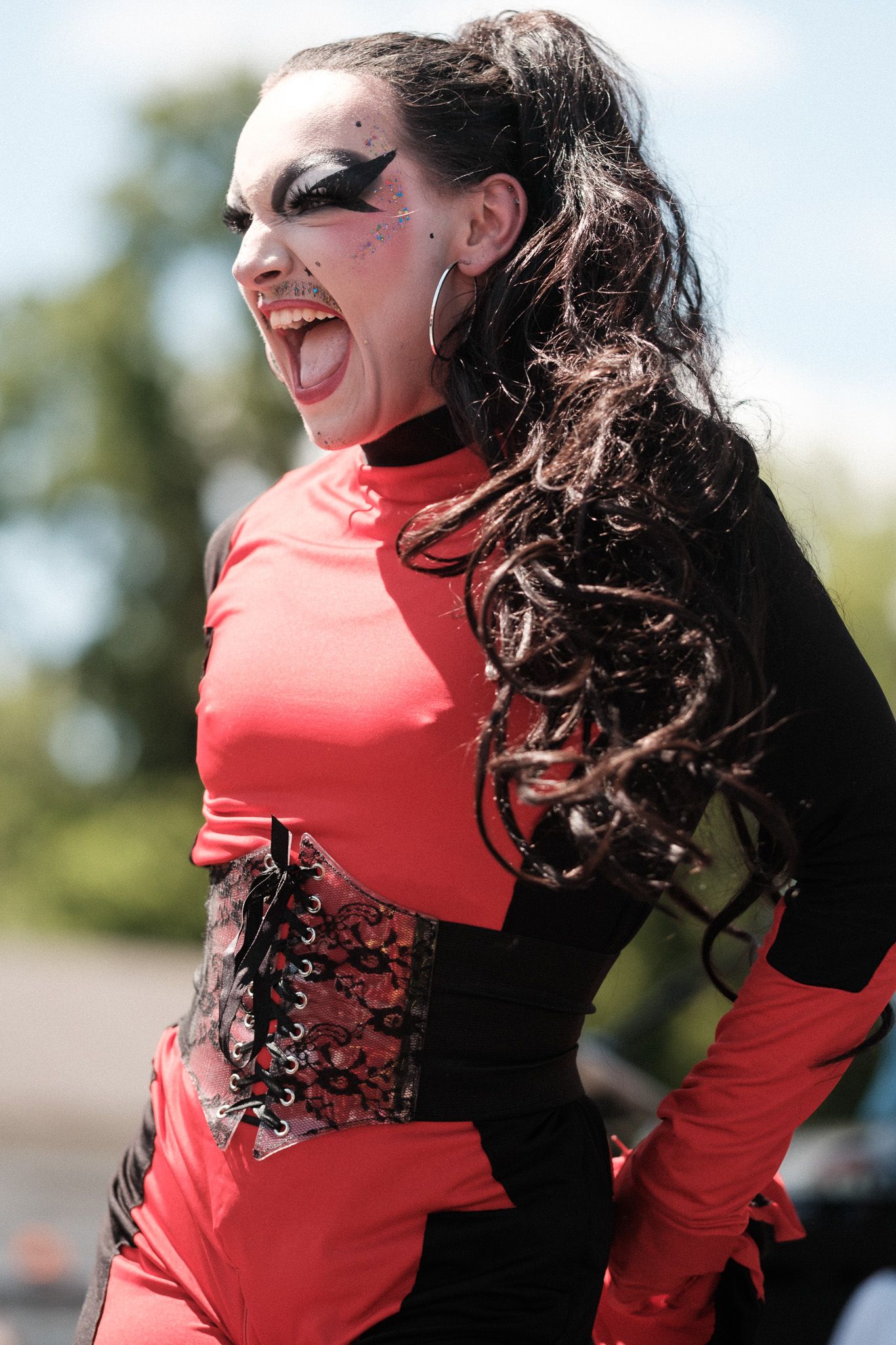 Dramatic performer in red and black costume with intricate makeup strikes an energetic theatrical pose outdoors.