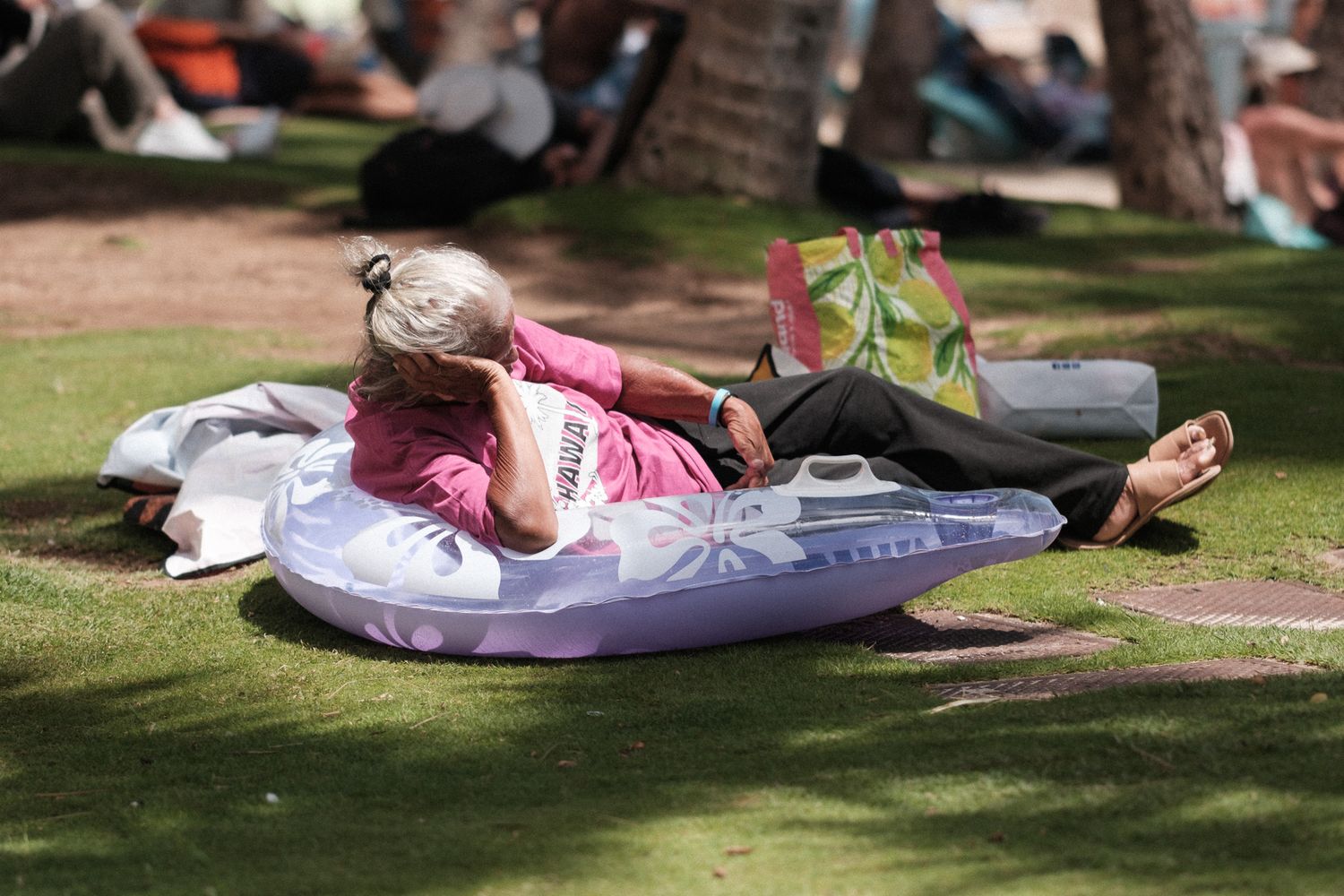 Someone relaxes on an inflatable air mattress while reading in a shaded park area on a warm day.