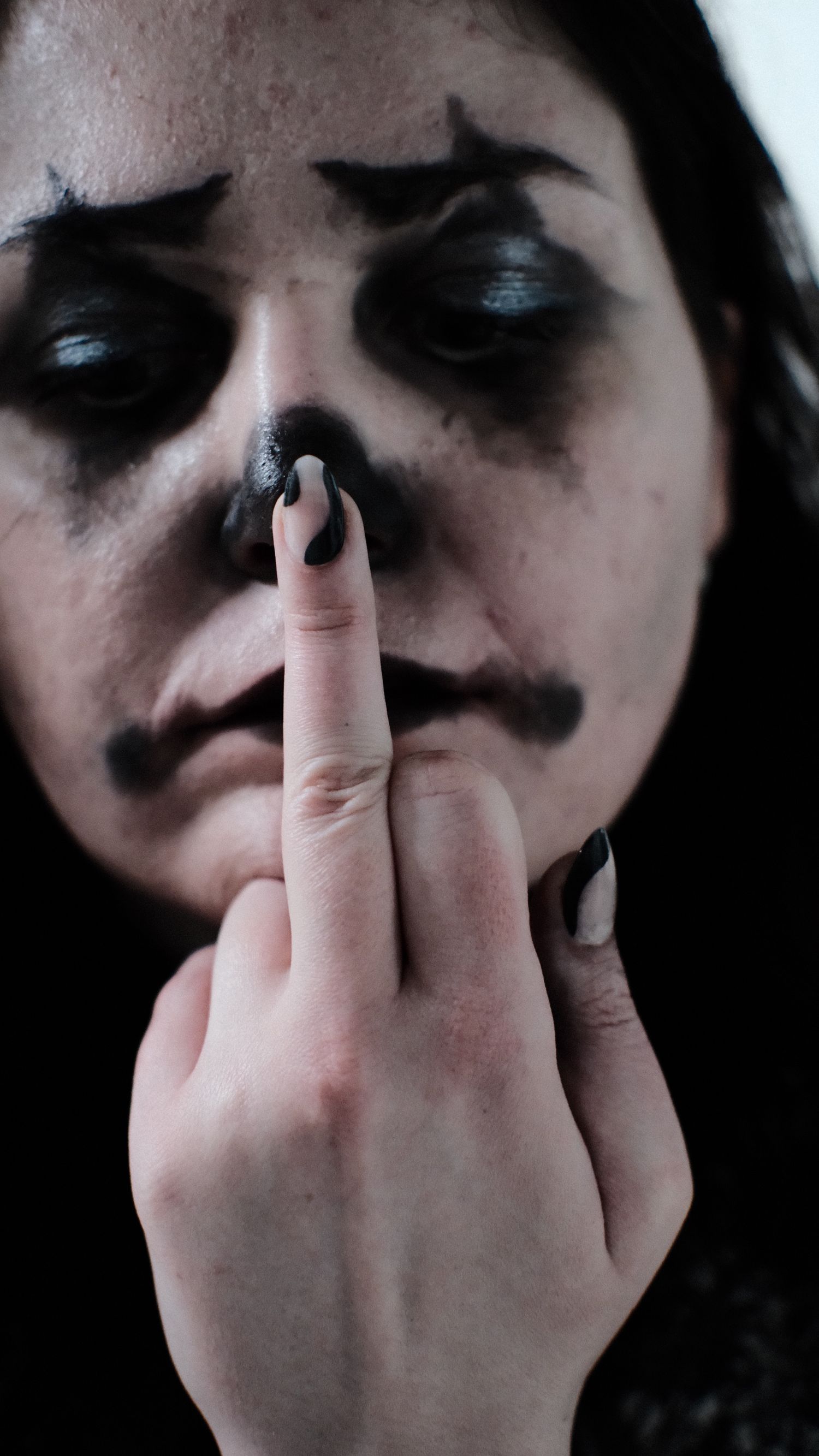 A person wearing dark gothic makeup holds up their middle finger to their nose in a dramatic portrait.