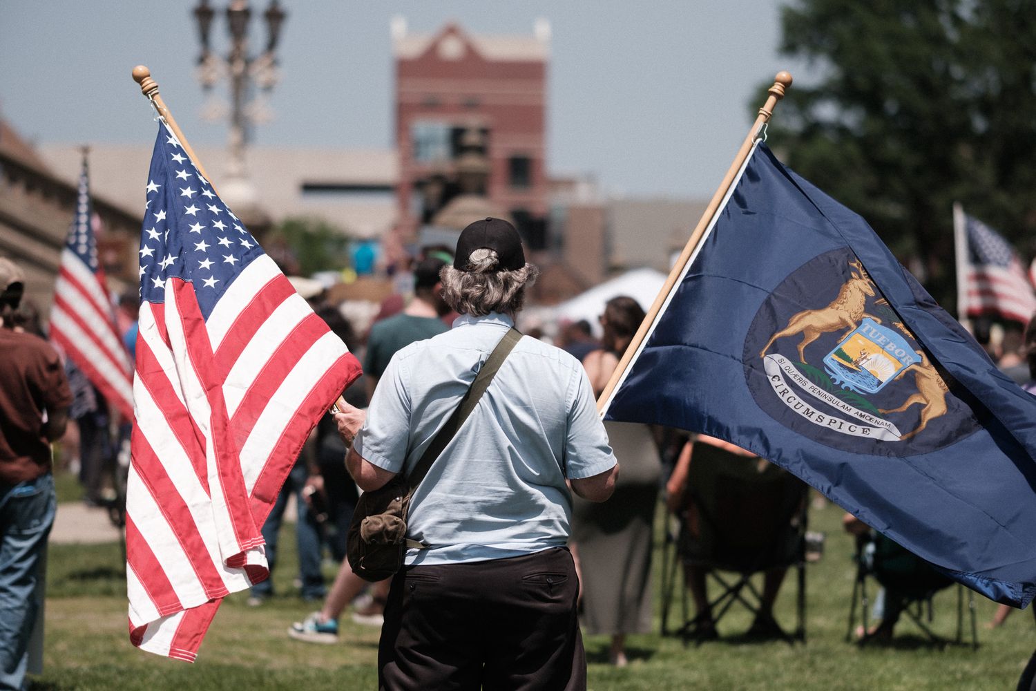 Person holds American flag and Michigan state flag at outdoor gathering with crowd and building in background.