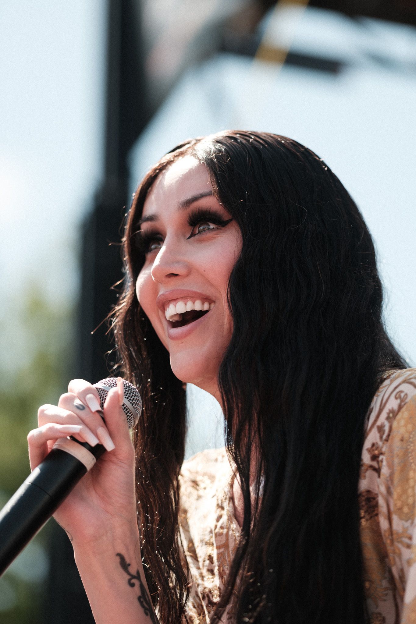 A performer with black hair sings passionately into a microphone on stage while wearing a gold-colored top.